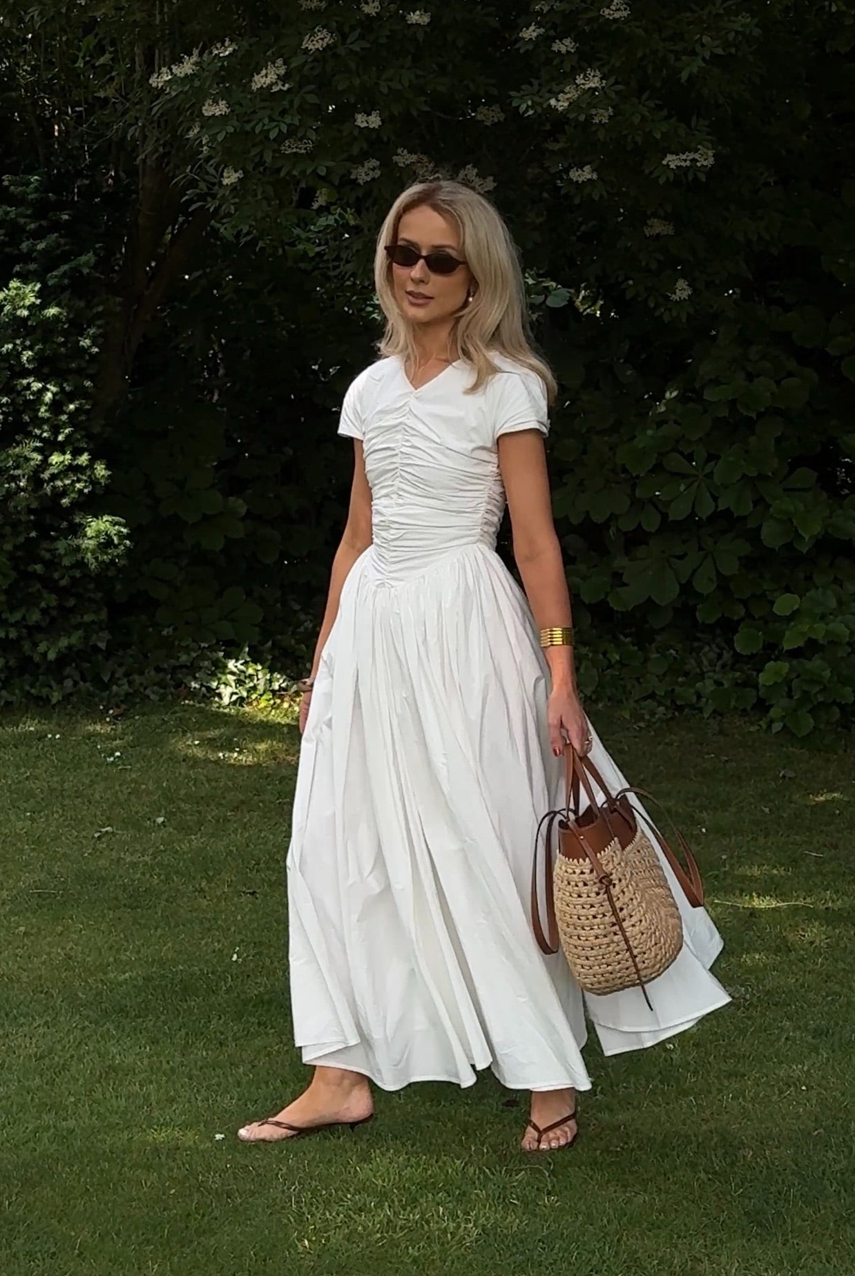 Model contrasting against an outdoor garden setting with a crisp white maxi dress featuring a v-neckline, short sleeves, full gathered fabric which is voluminous and flowing, styled with sunglasses, sandals and a straw tote