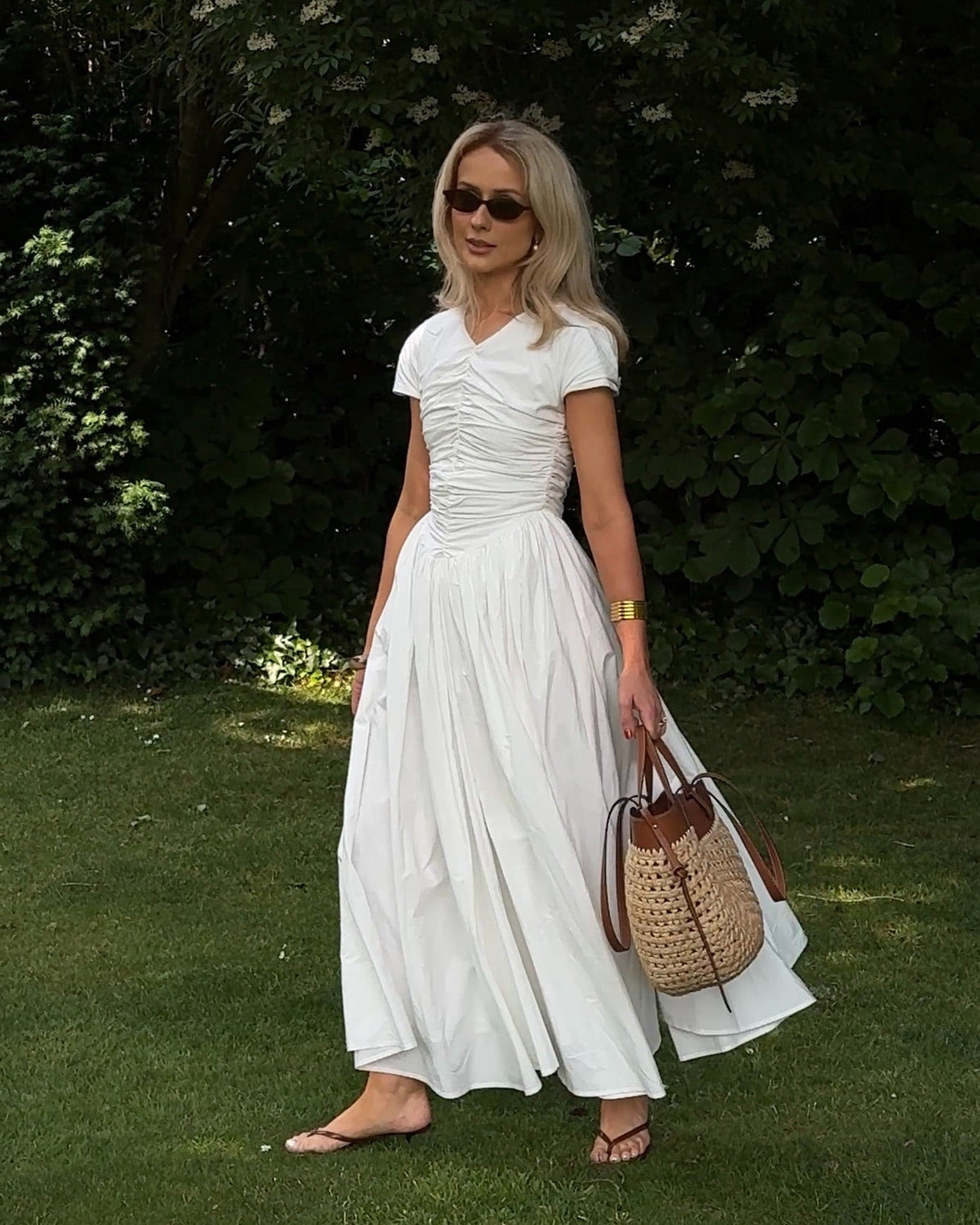 Model contrasting against an outdoor garden setting with a crisp white maxi dress featuring a v-neckline, short sleeves, full gathered fabric which is voluminous and flowing, styled with sunglasses, sandals and a straw tote