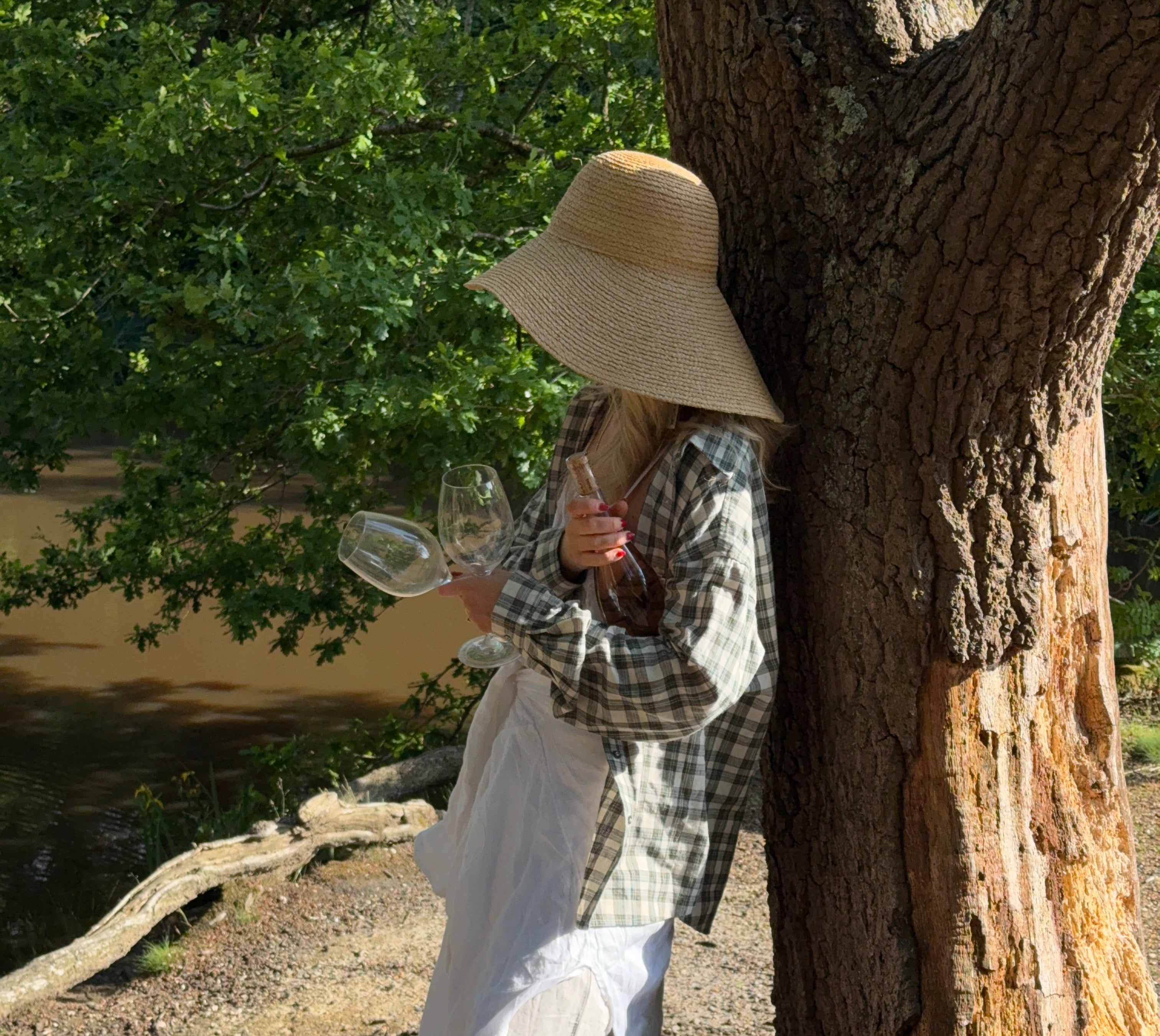 Person standing under a large tree while holding a bottle of wine and two glasses, wearing a checkered shirt and white outfit