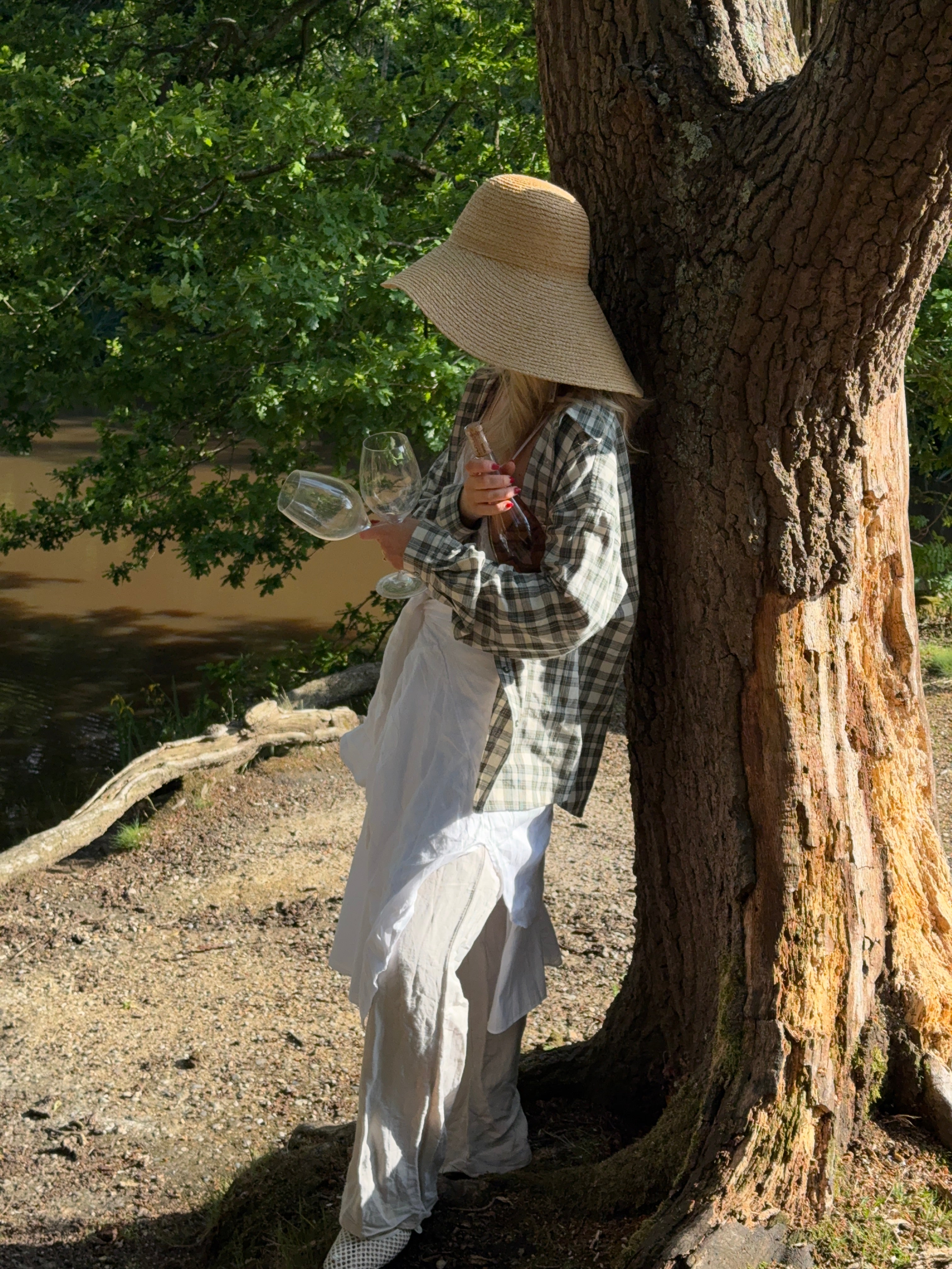 Person standing under a large tree while holding a bottle of wine and two glasses, wearing a checkered shirt and white outfit