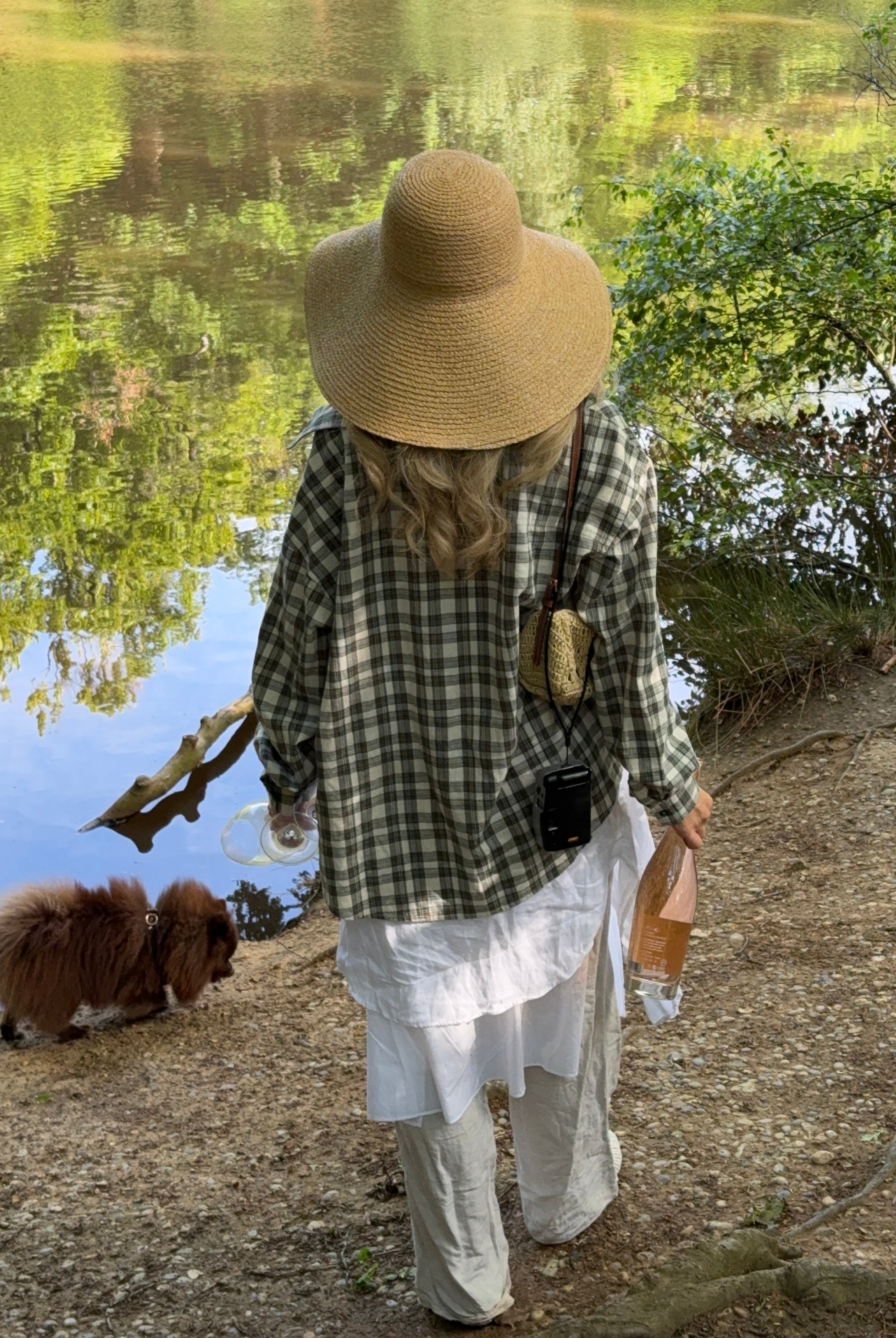 Model walking a dog by a lake, wearing a checkered shirt and wide-brimmed hat holding an unopened wine bottle