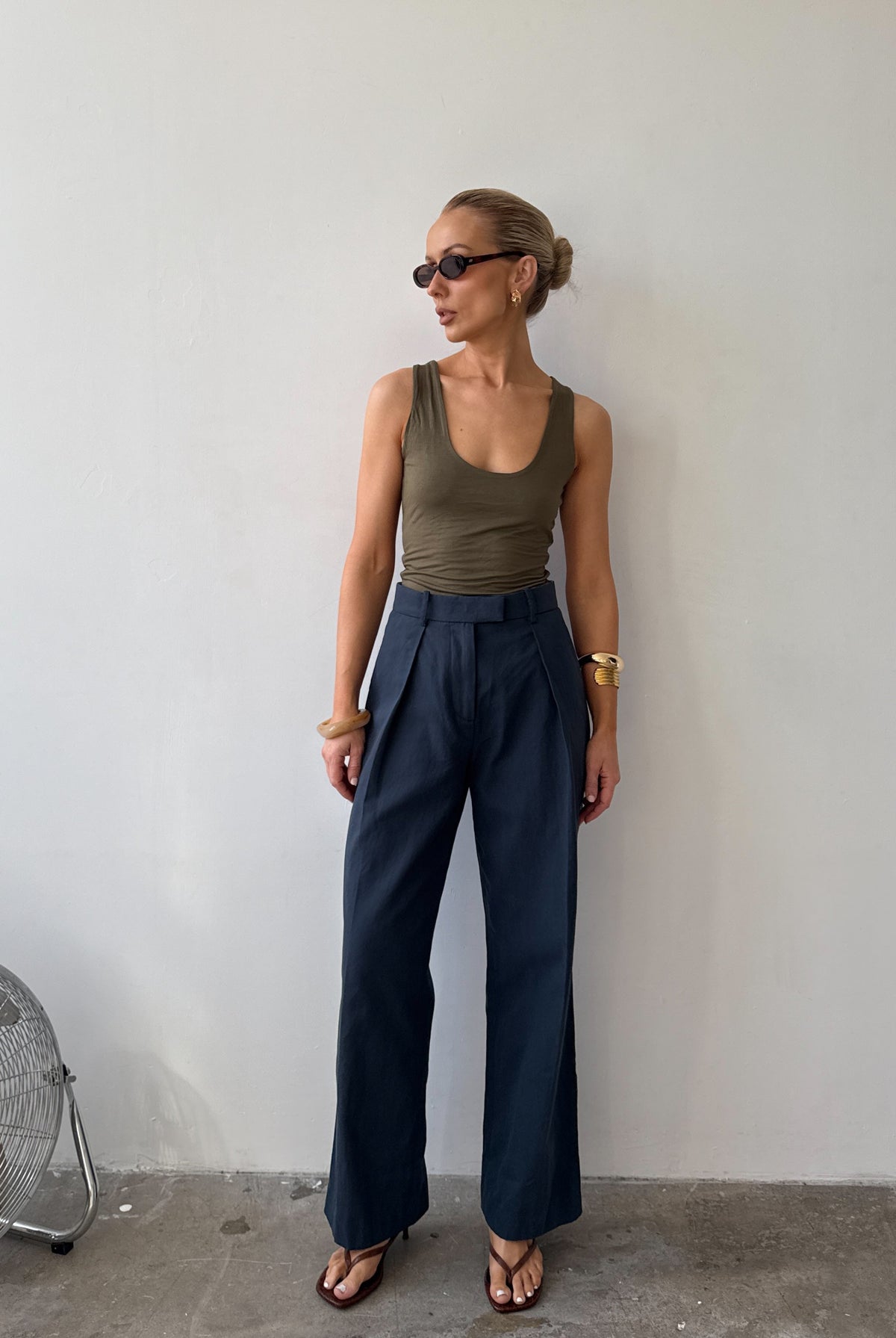 Model wearing ink blue tailored trousers with front pleats styled with a green tank top, with sunglasses on and flat sandals.