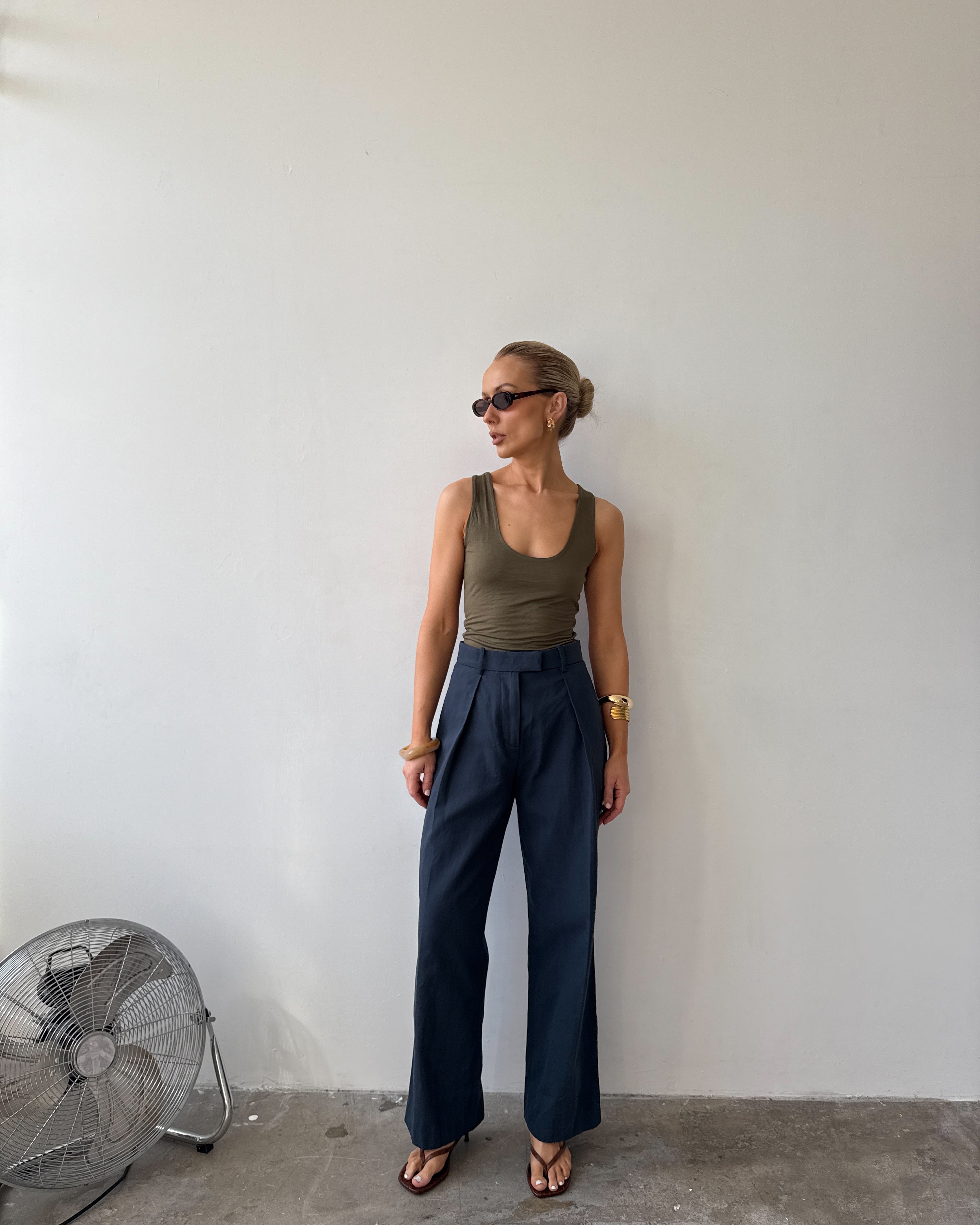 Model wearing ink blue tailored trousers with front pleats styled with a green tank top, with sunglasses on and flat sandals.