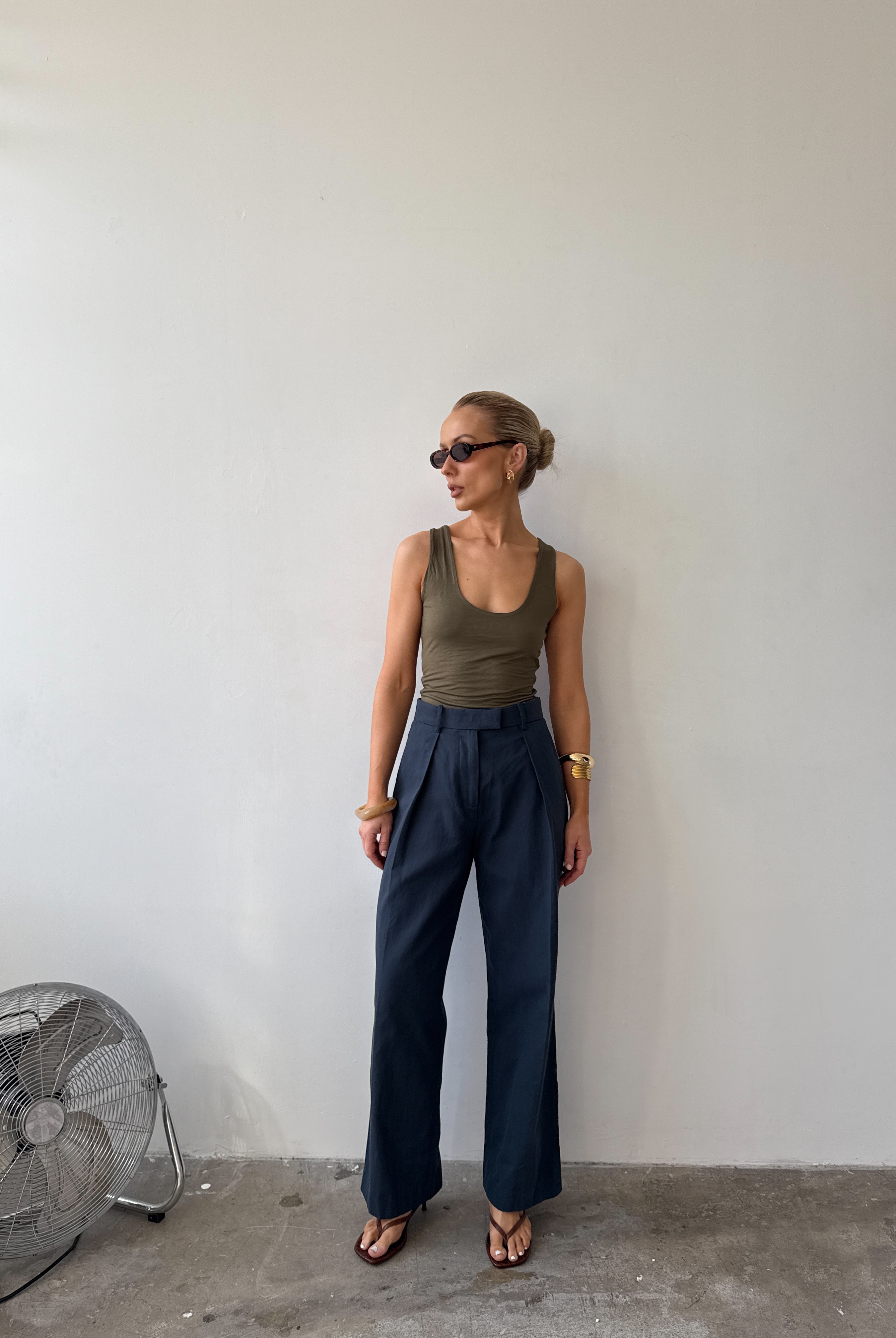 Model wearing ink blue tailored trousers with front pleats styled with a green tank top, with sunglasses on and flat sandals.