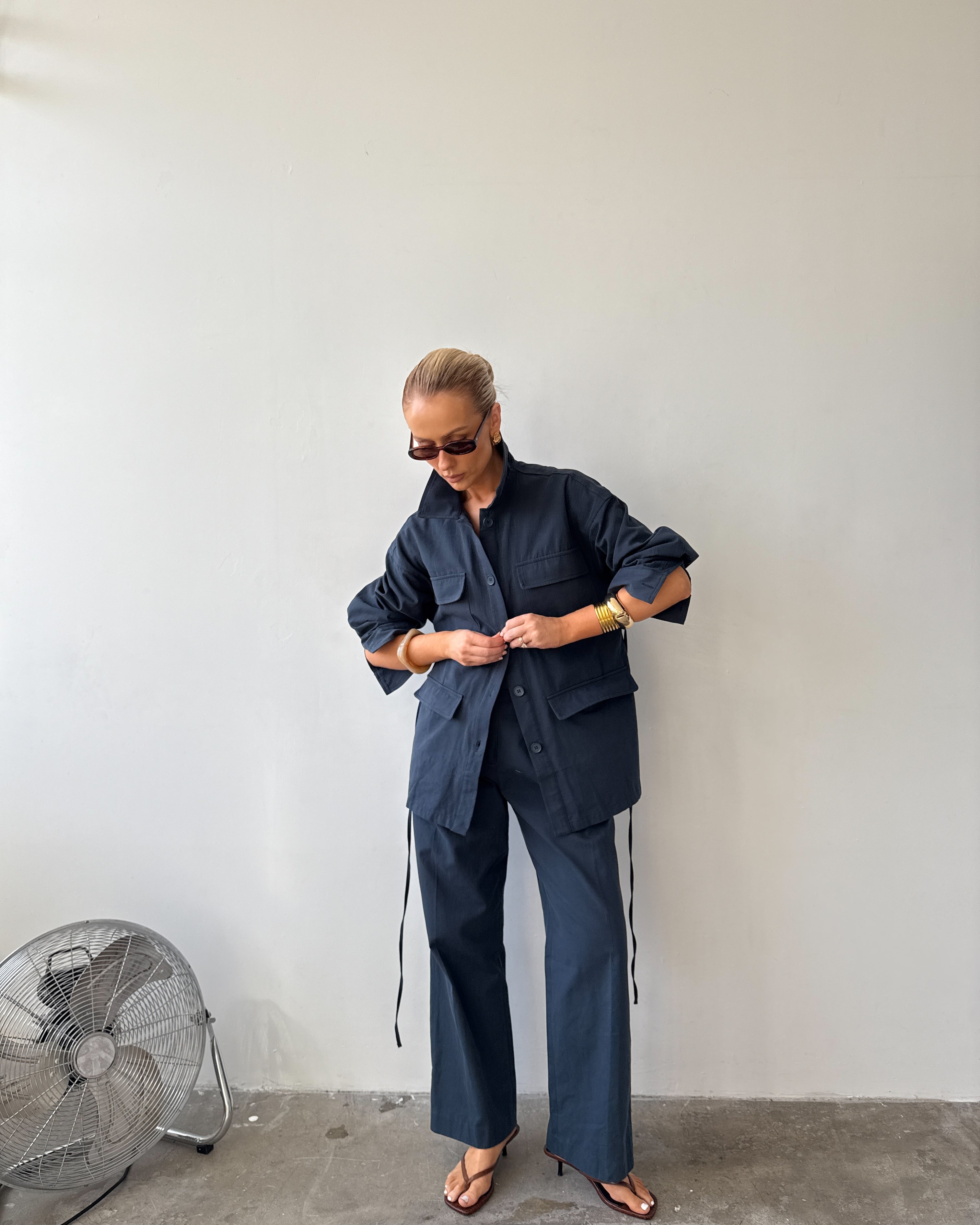 Model wearing navy two‑piece set (The Ink Blue Field Jacket and Day Trousers) while standing against a plain pale wall, wearing sunglasses, gold bracelets and sandals.
