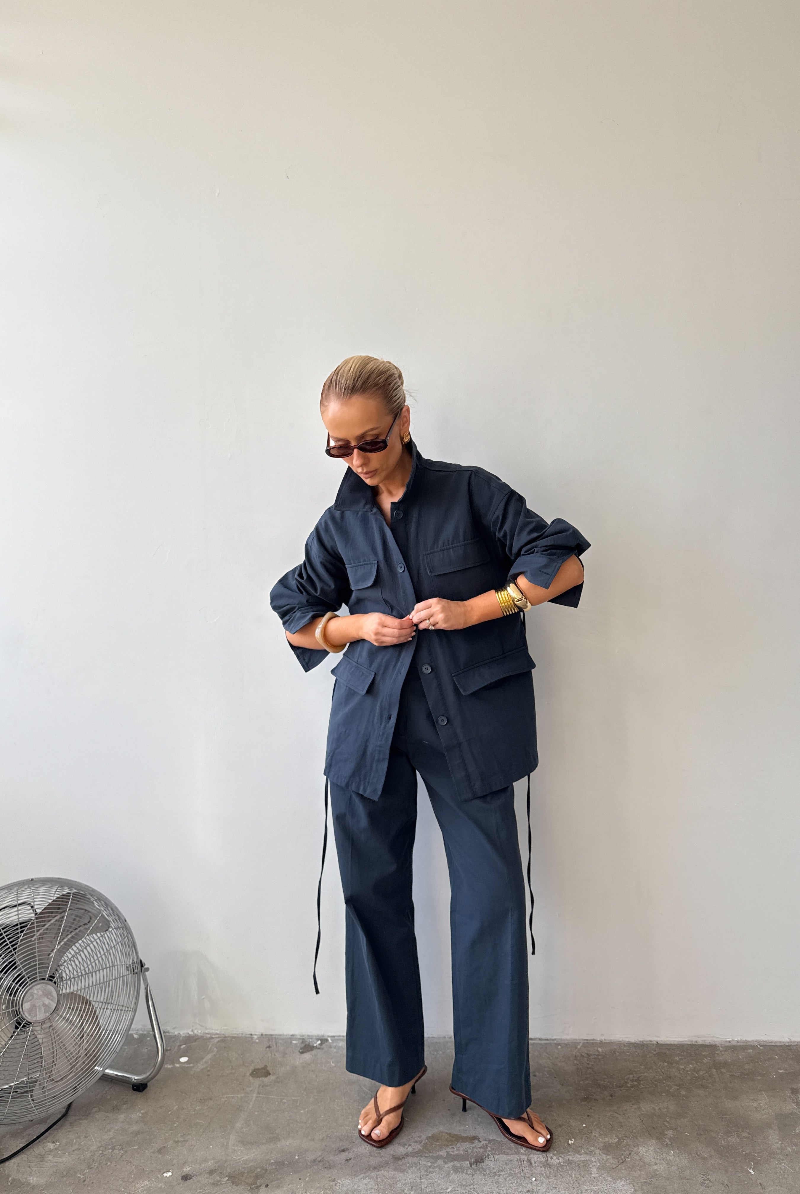 Model wearing navy two‑piece set (The Ink Blue Field Jacket and Day Trousers) while standing against a plain pale wall, wearing sunglasses, gold bracelets and sandals.