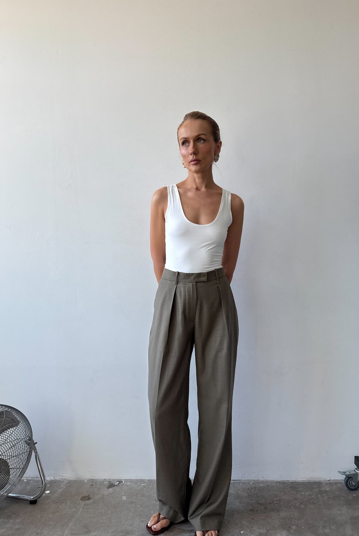 Model wearing a white tank top and light neutral green wide-leg pants with sandals standing against a plain wall.