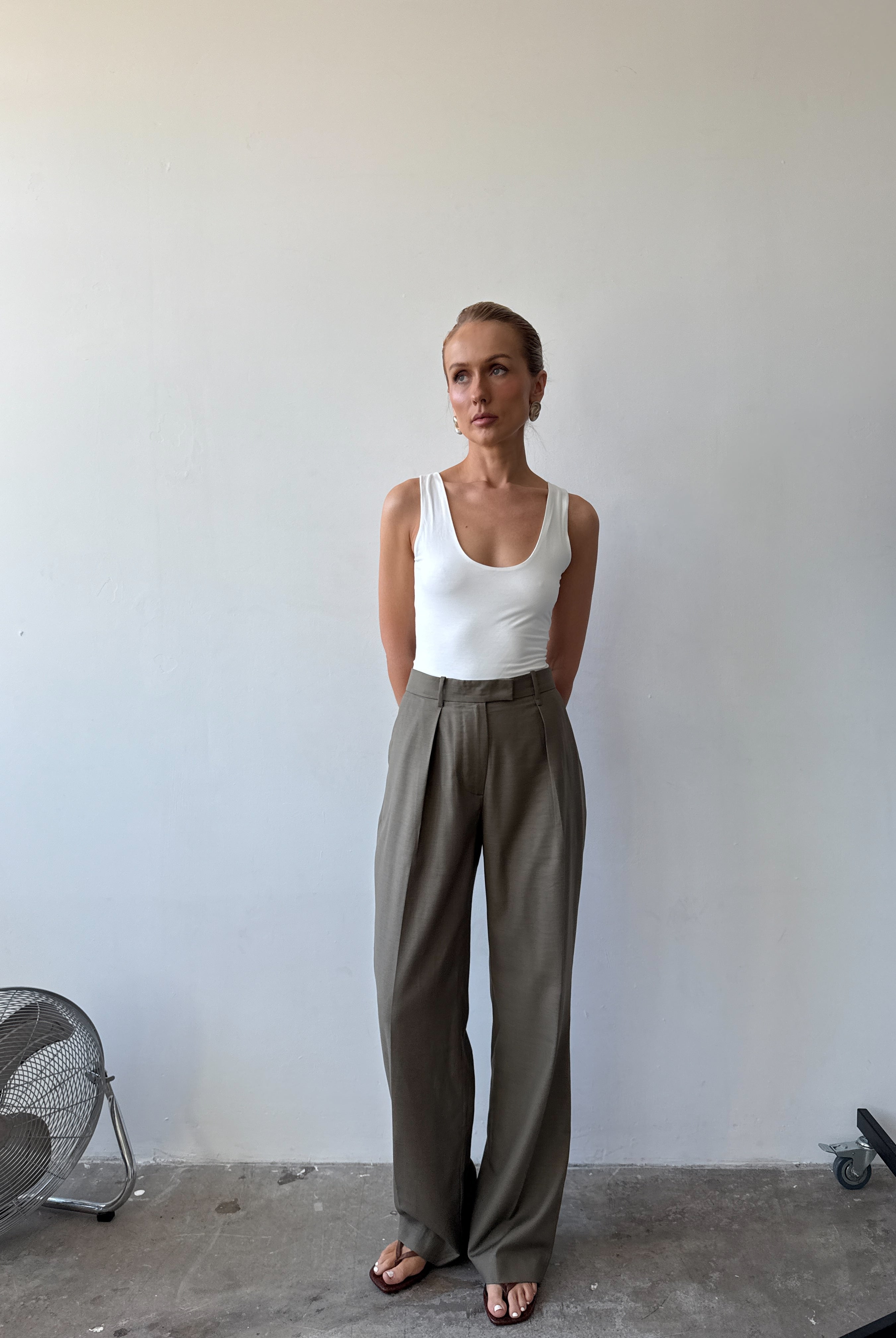 Model wearing a white tank top and light neutral green wide-leg pants with sandals standing against a plain wall.