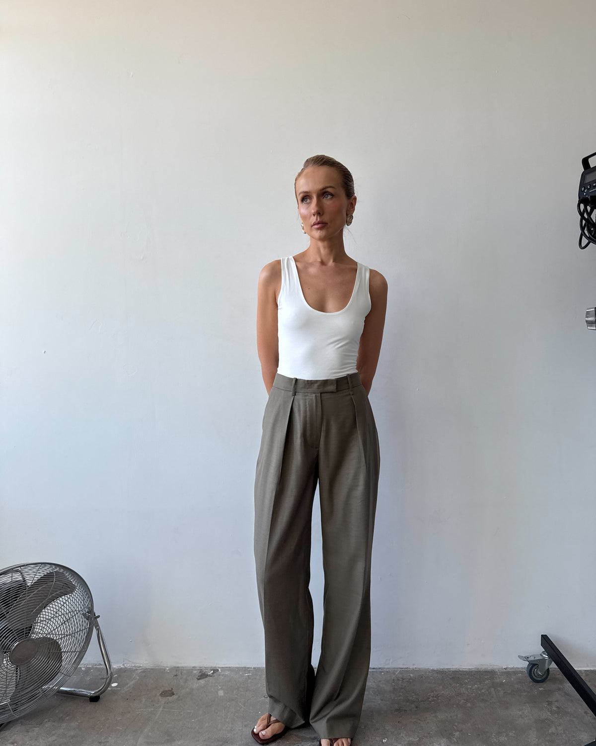 Model wearing a white tank top and light neutral green wide-leg pants with sandals standing against a plain wall.