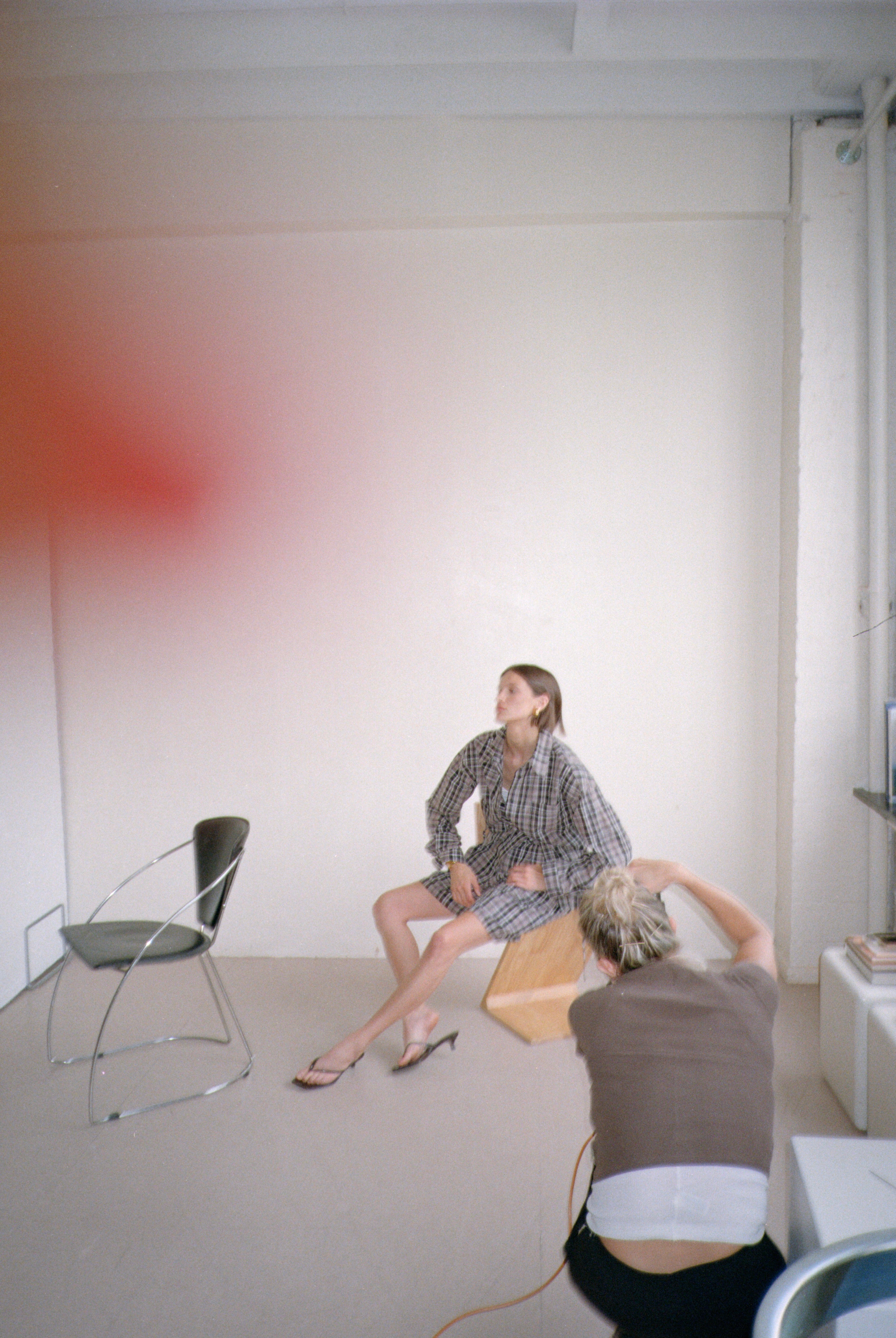 Two people in a room with minimal furniture, one photographs while the other poses wearing a plaid outfit