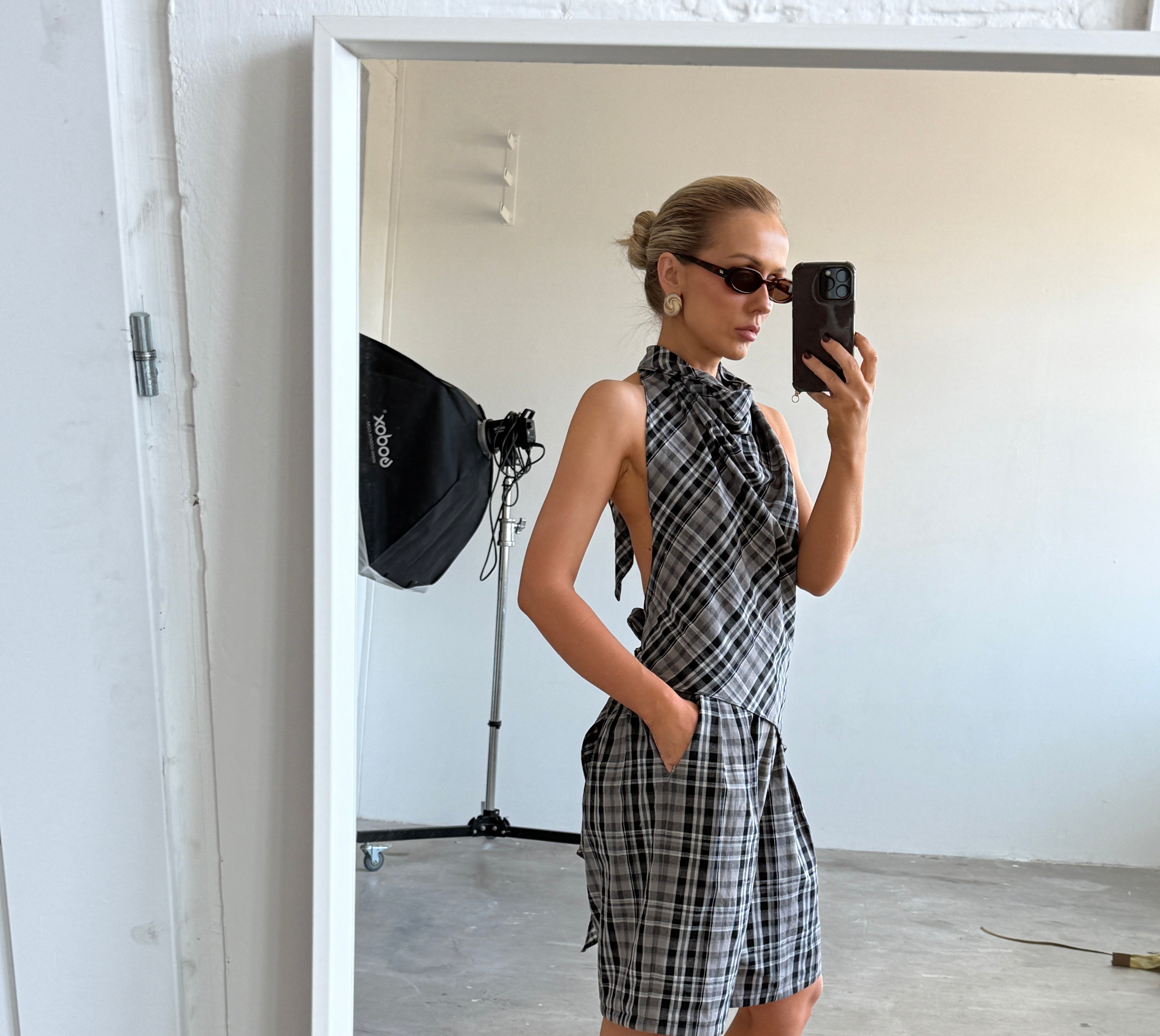Model taking a mirror selfie wearing a plaid backless top with matching shorts accessorised by sunglasses and heeled sandals in a minimalistic room