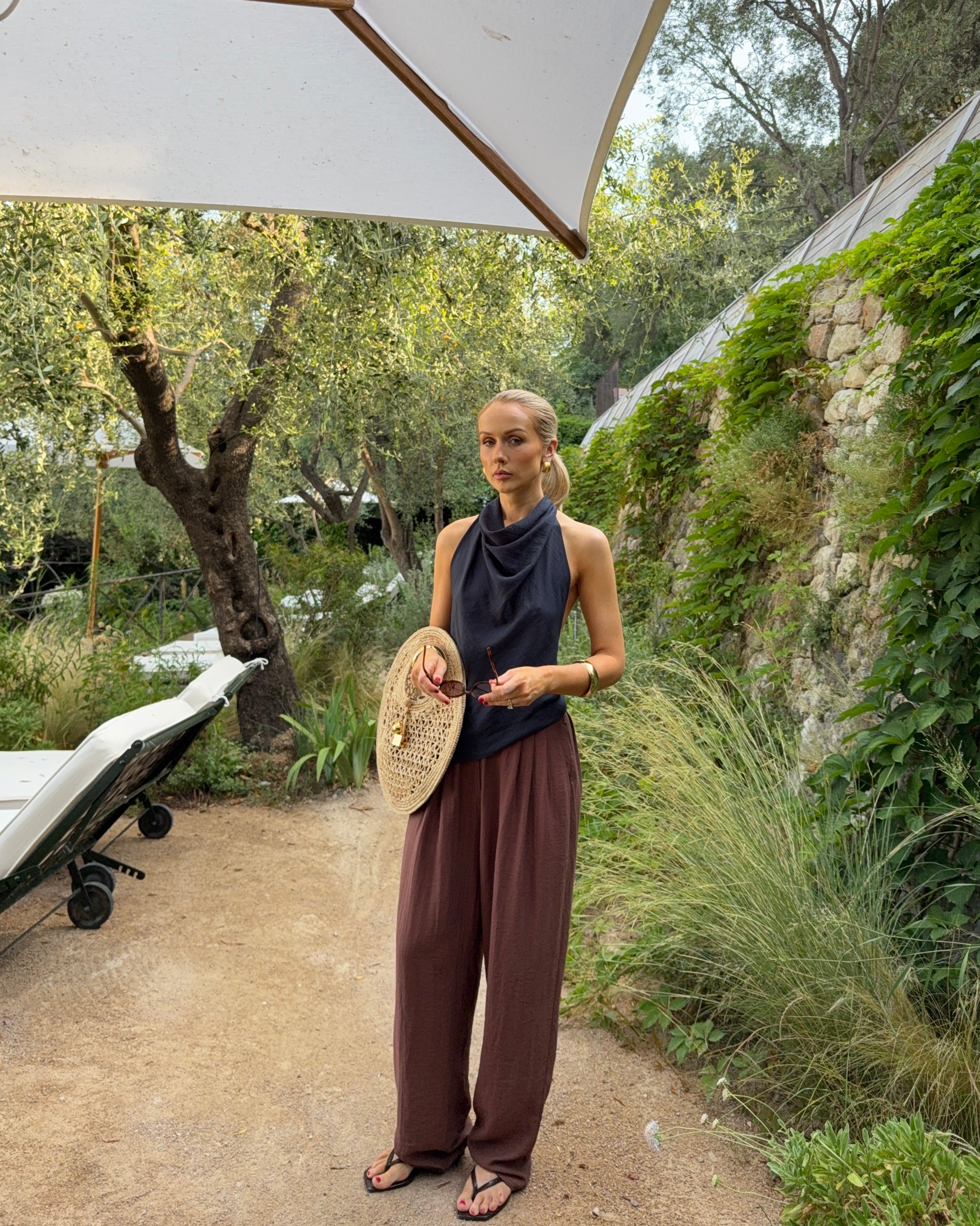 Model wearing a simple draping black top and brown linen trousers while holding a statement bag in a natural holiday setting with greenery and a stone wall.