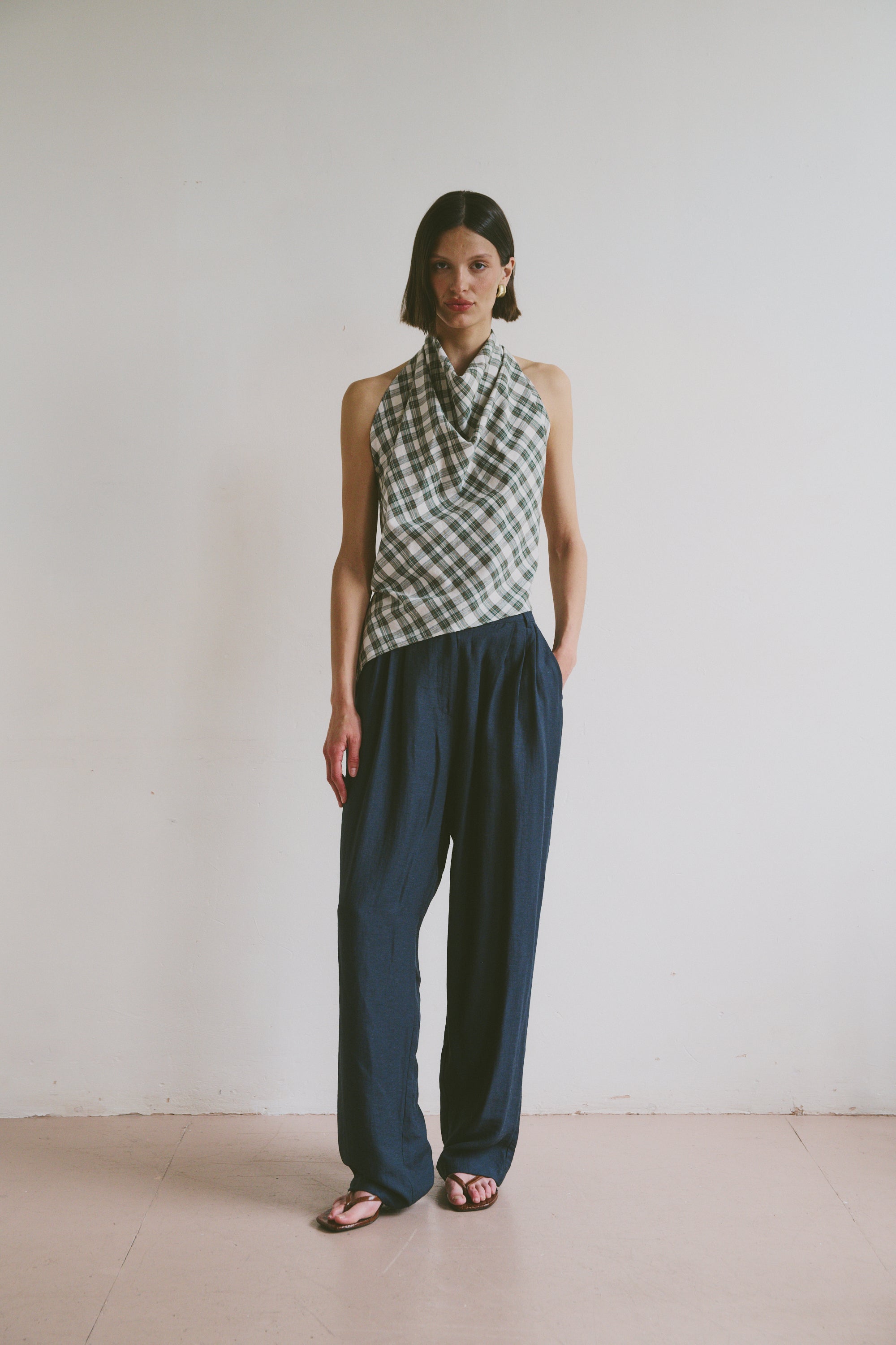Model wears ink blue, flowy linen trousers styled with a checkered, high-neck sleeveless top and black sandals