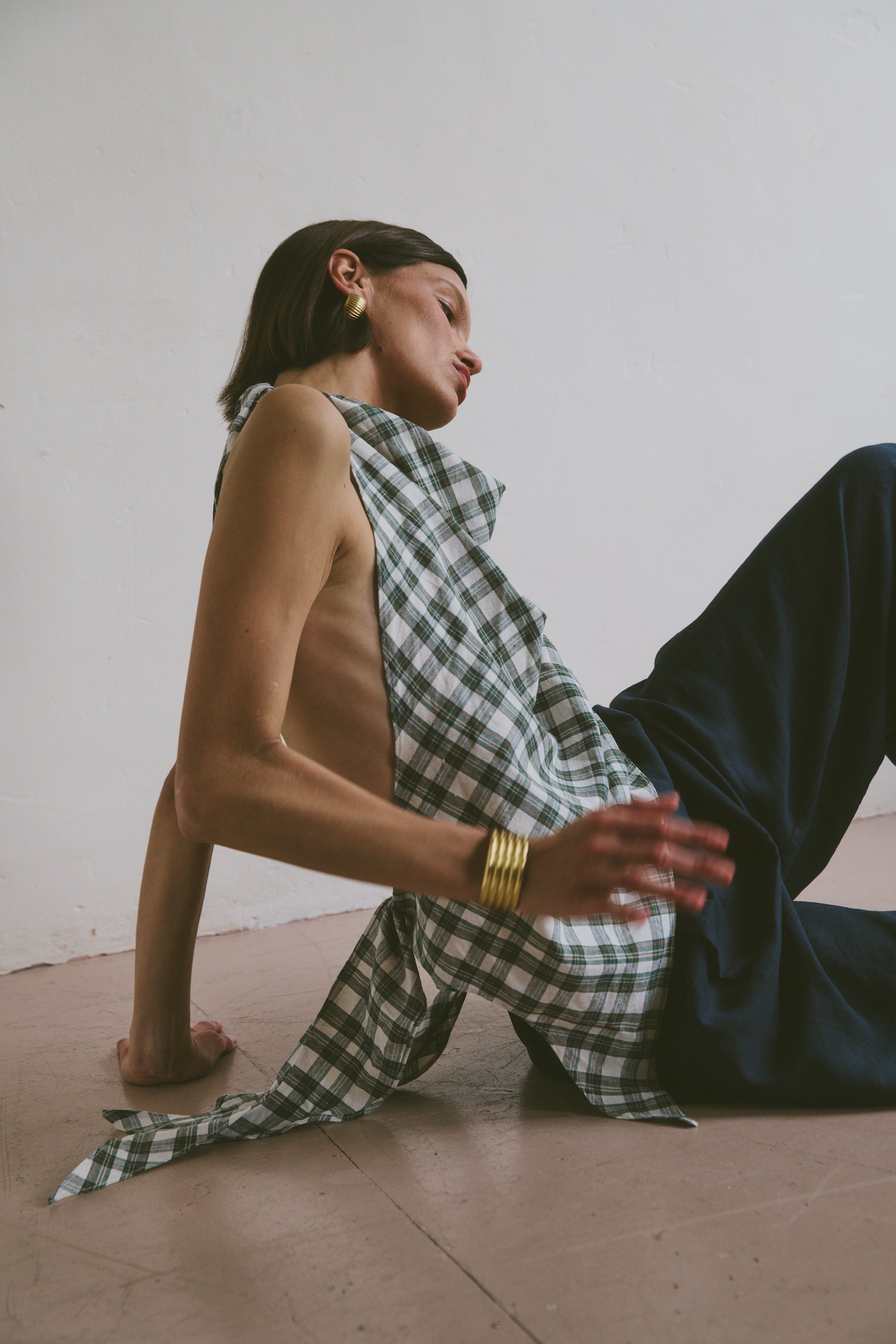 Model wears a draping checkered top with an open back tied at the bottom, paired with dark blue trousers