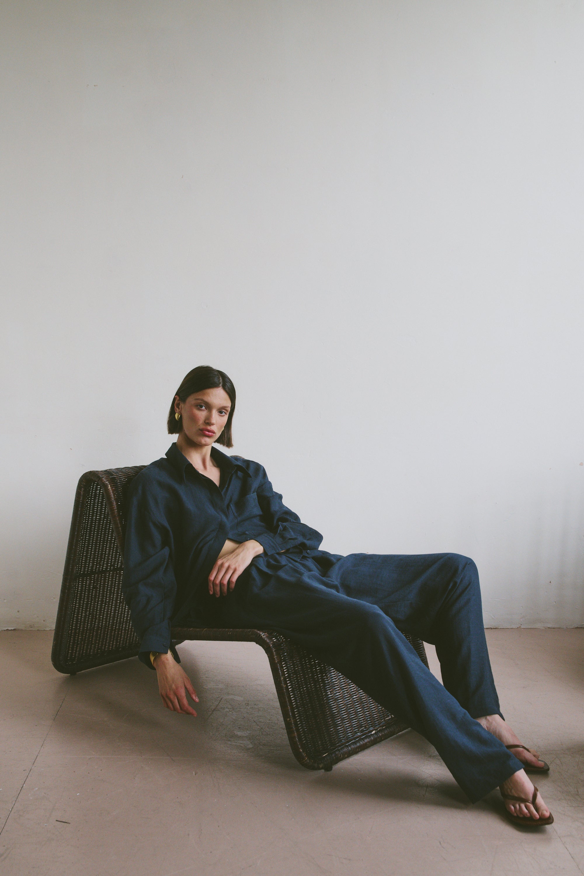 Model in a navy blue outfit sitting on a wicker chair in a relaxed position against a plain wall