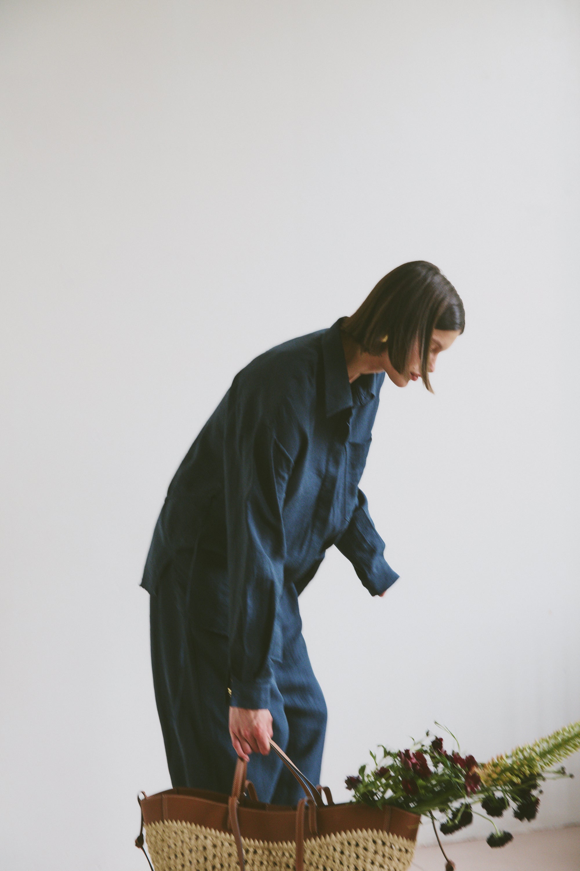 Model wearing a blue outfit holding a woven handbag with flowers and greenery coming out the top 