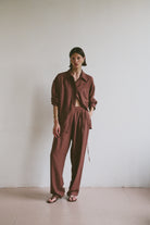 Model wearing a relaxed maroon linen shirt and matching trousers styled with open toe sandals in front of a minimalistic background