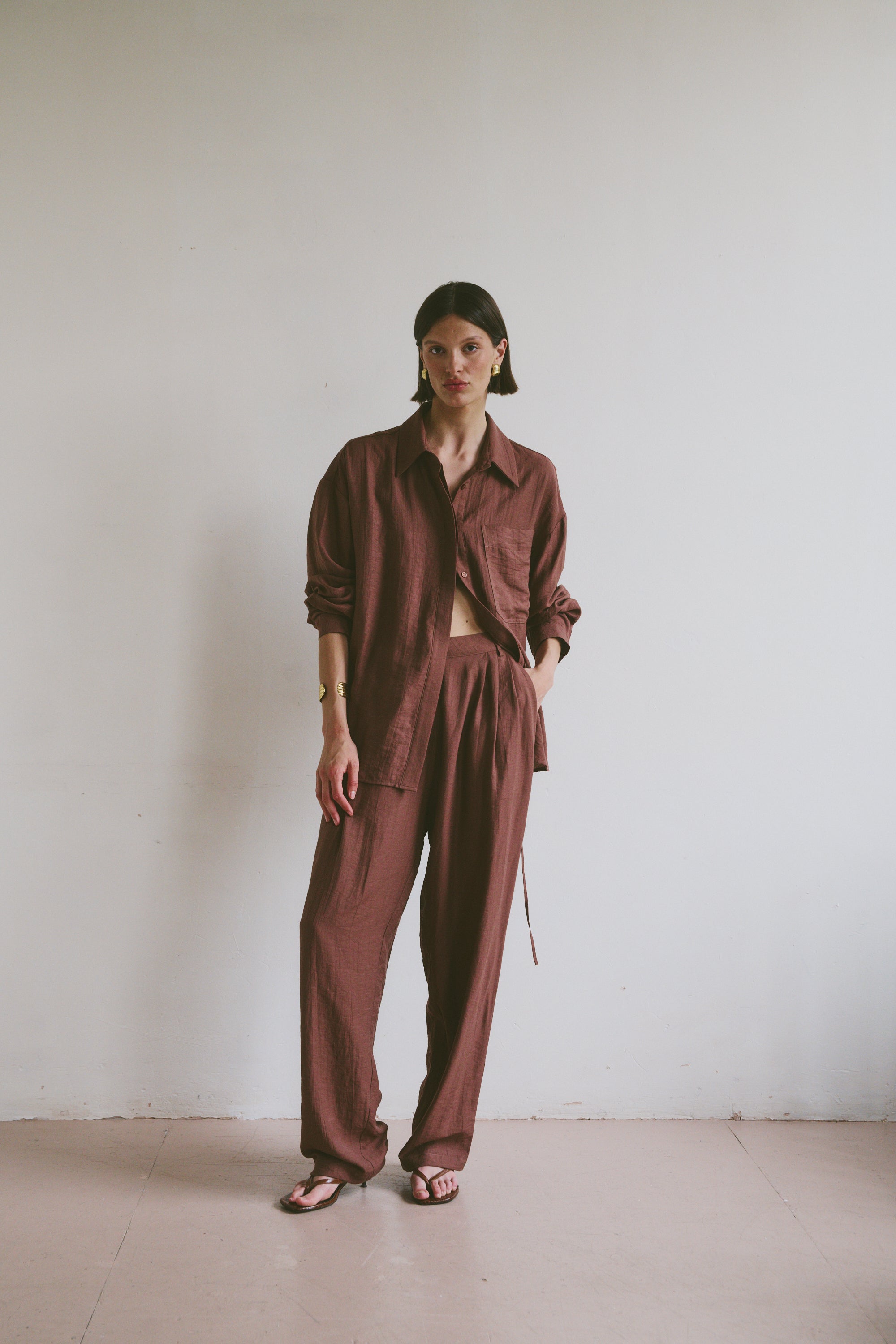 Model wearing a relaxed maroon linen shirt and matching trousers styled with open toe sandals in front of a minimalistic background