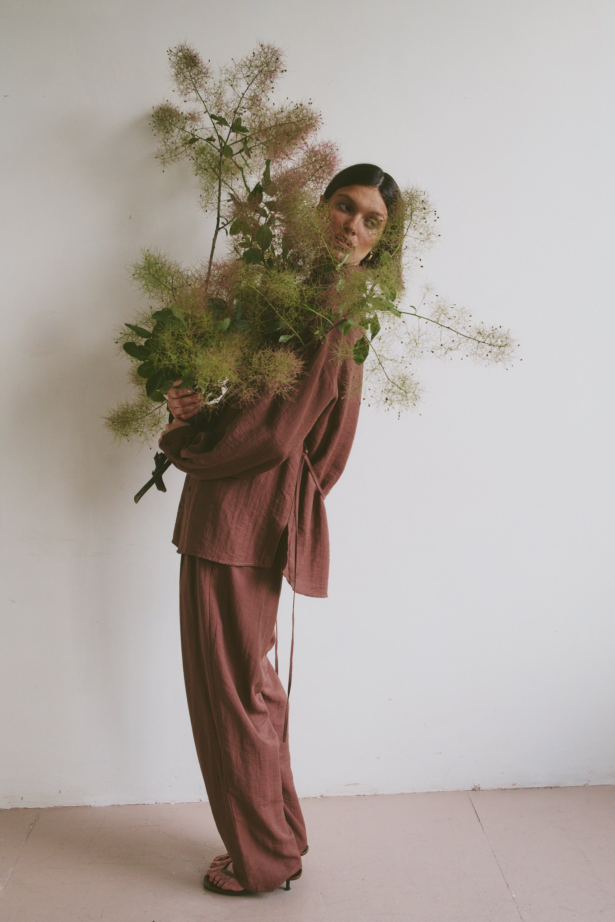 Model in a loose-fitting, mauve-toned outfit holding a large, airy bouquet of greenery, standing against a plain white wall