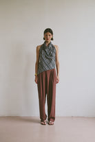 Model wearing a patterned top tied at the neck and maroon brown trousers against a plain background