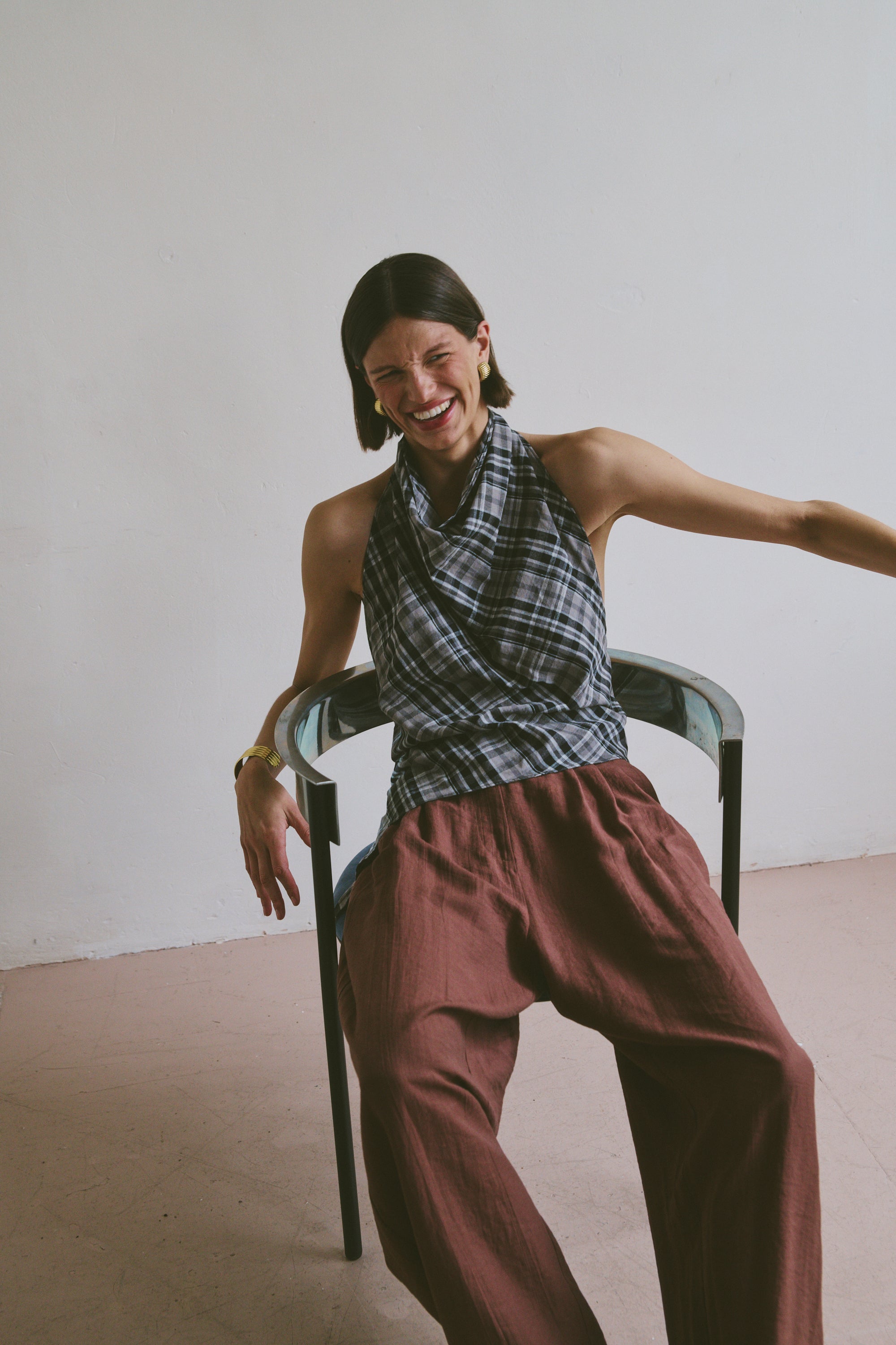 Woman laughing while sitting on a chair wearing a plaid top and burgundy pants against a plain background