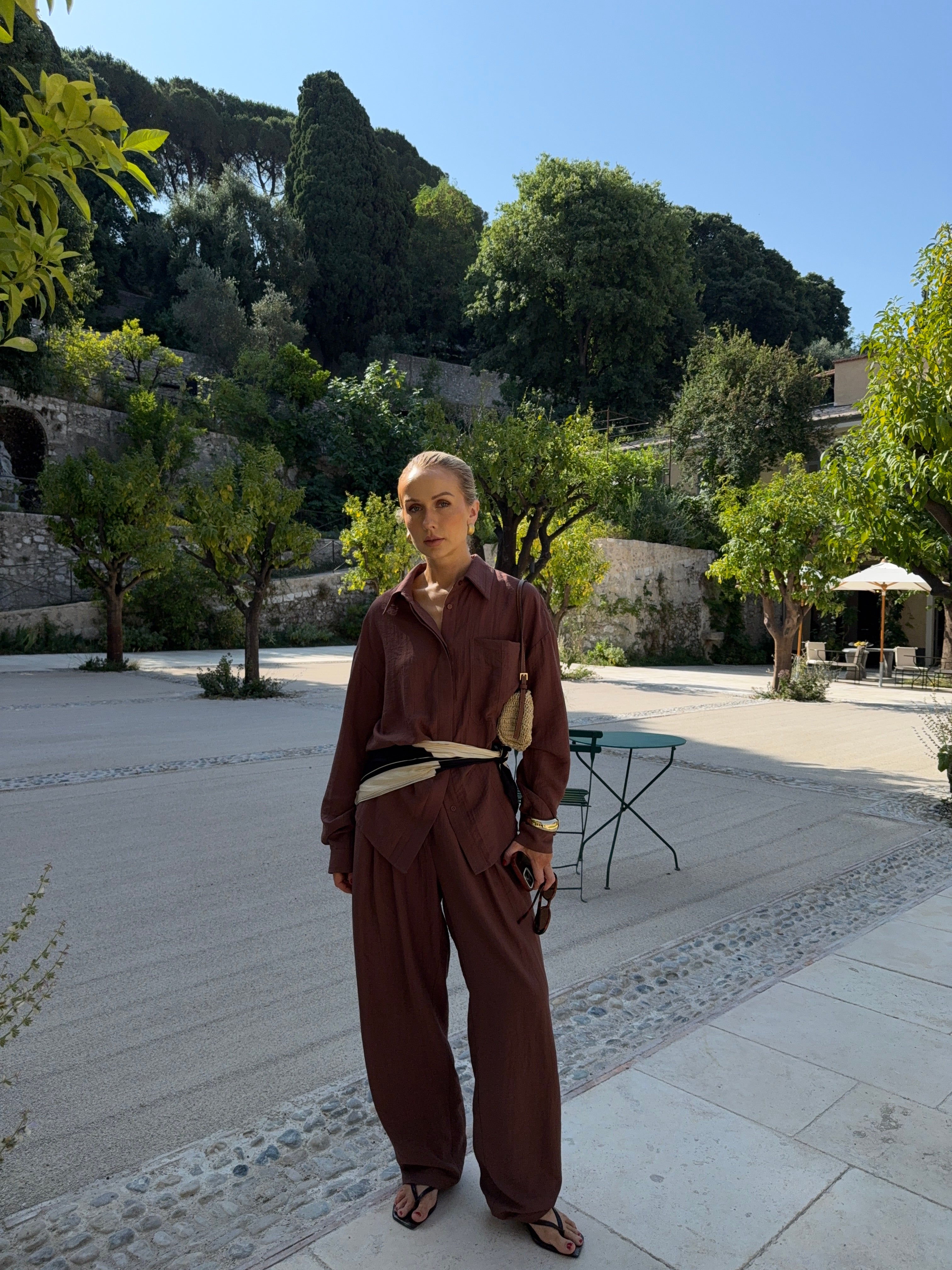 Woman in auburn-brown outfit standing on a paved area with trees and buildings in the background