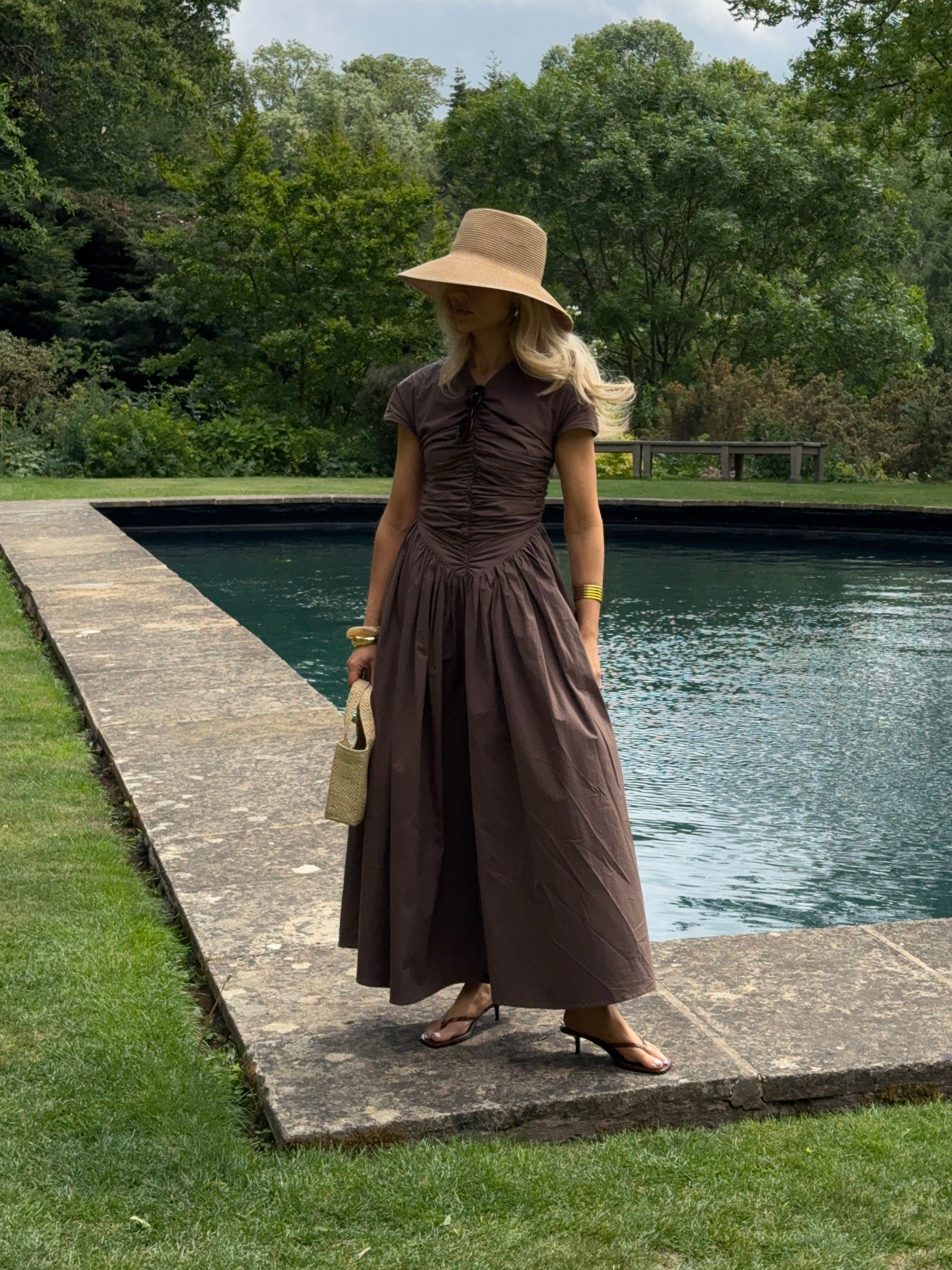 The model wears a brown, short-sleeved ruched dress with a full, flowing ankle-length skirt. She styles the look with a wide-brimmed straw hat, gold bangles, and a small textured handbag, striking a graceful pose in front of a rectangular reflecting pool bordered by a stone walkway and surrounded by lush greenery