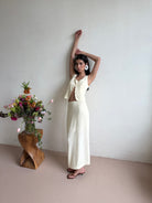 Model in a yellow-white outfit standing next to a wooden stool with flowers against a plain wall