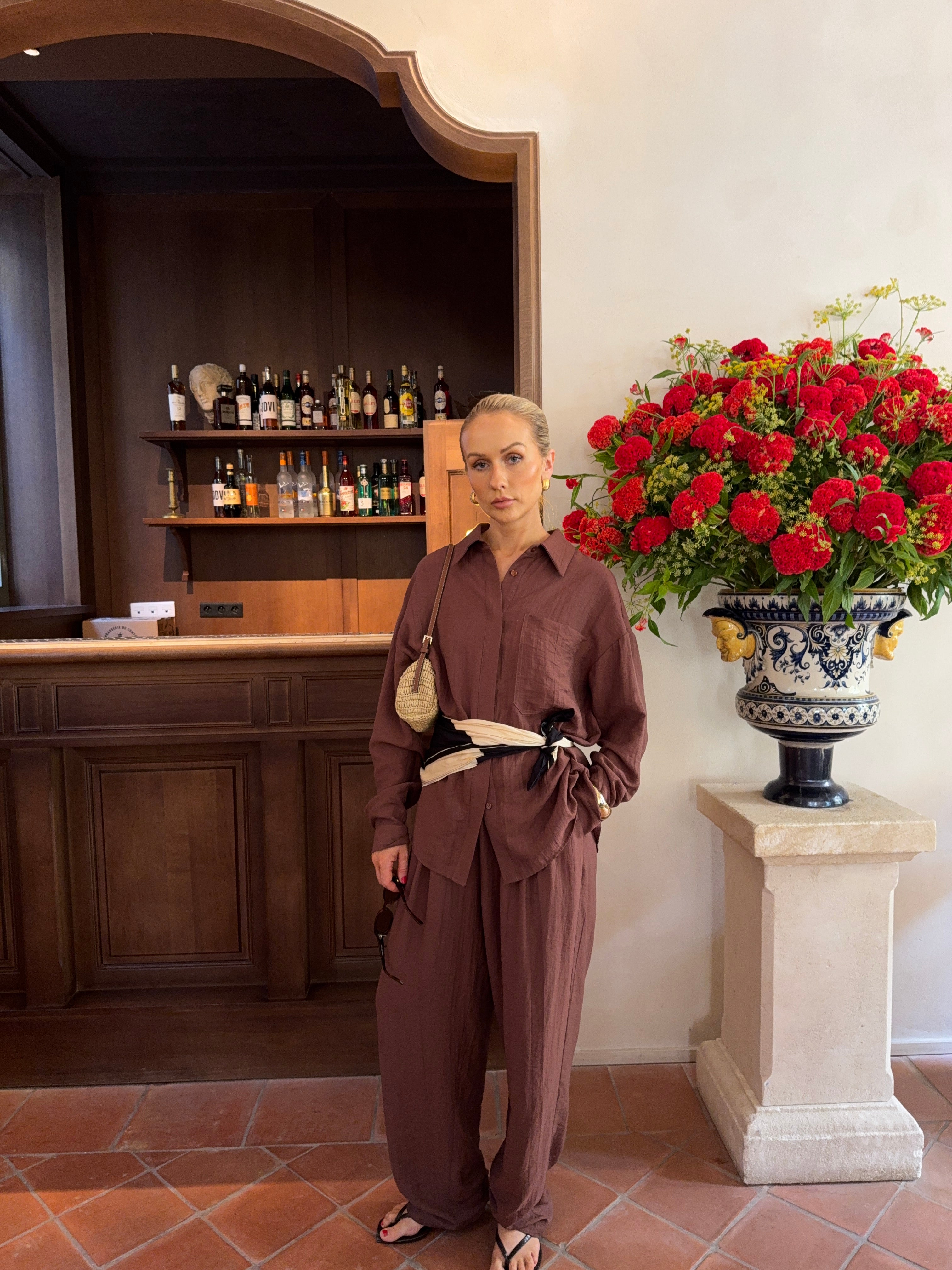 Model in a brown linen shirt and matching trousers accessorised with a scarf bel and handbag, standing in front of a large red and yellow floral arrangement and wooden bar