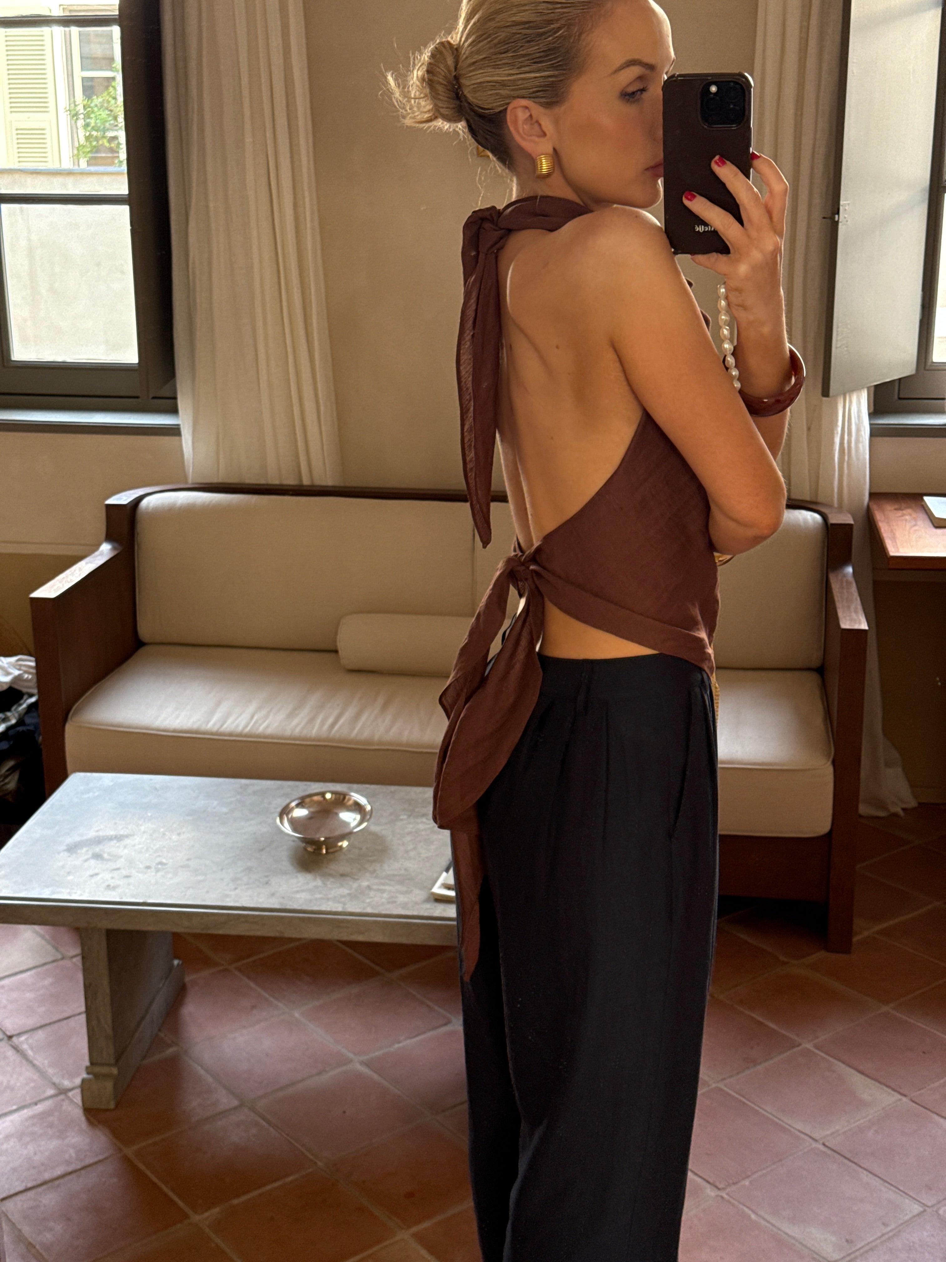 Person taking a mirror selfie in a room with a couch and window while wearing a backless top thats tied at the top and bottom with black trousers
