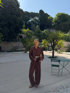 Model in a brown outfit standing on a paved area with a Mediterranean backdrop of trees and stone walls