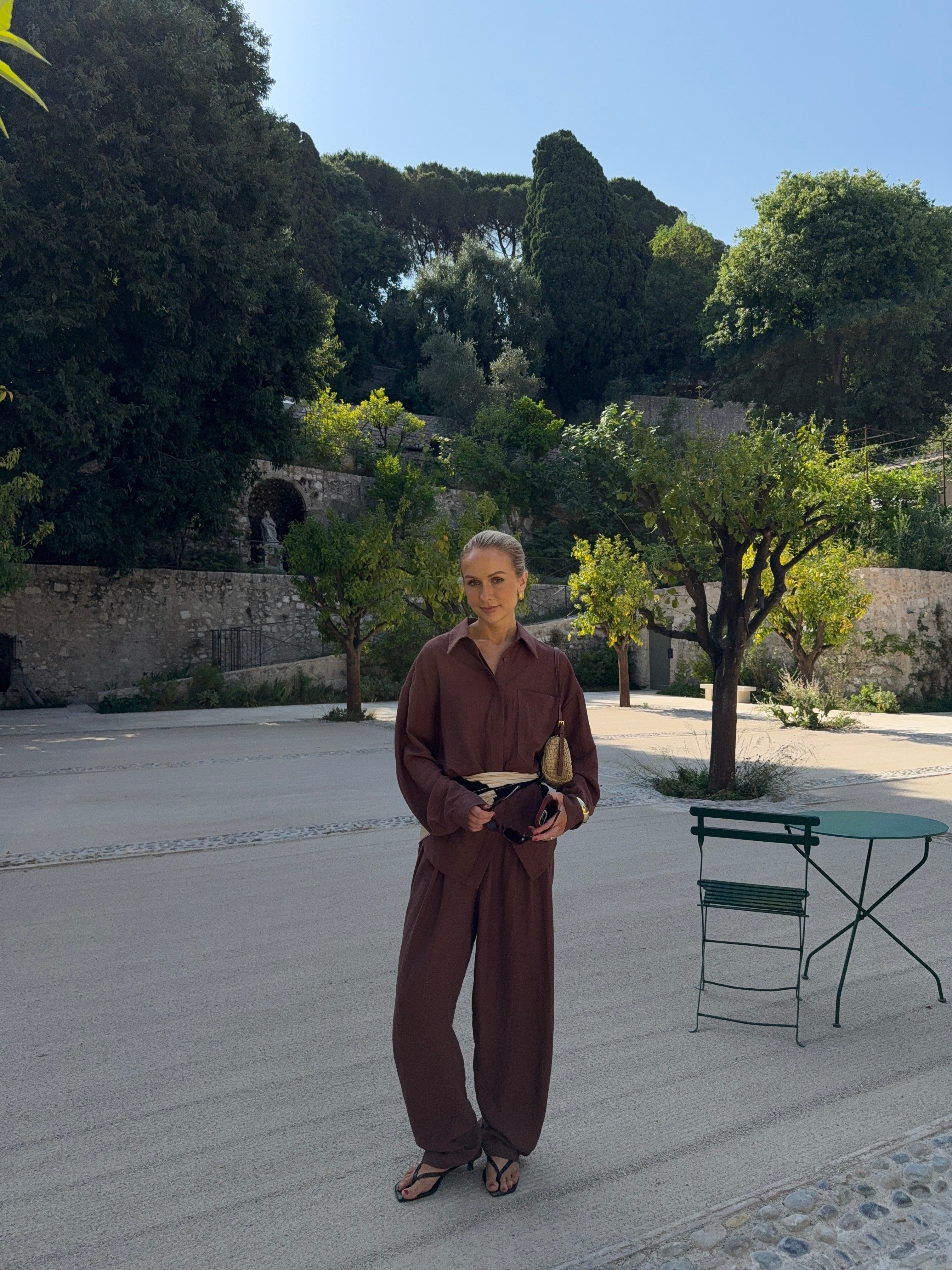 Model in a brown outfit standing on a paved area with a Mediterranean backdrop of trees and stone walls