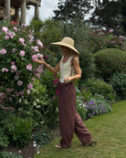 Woman in a vibrant flower garden wearing a wide-brimmed hat and flowing maroon trousers