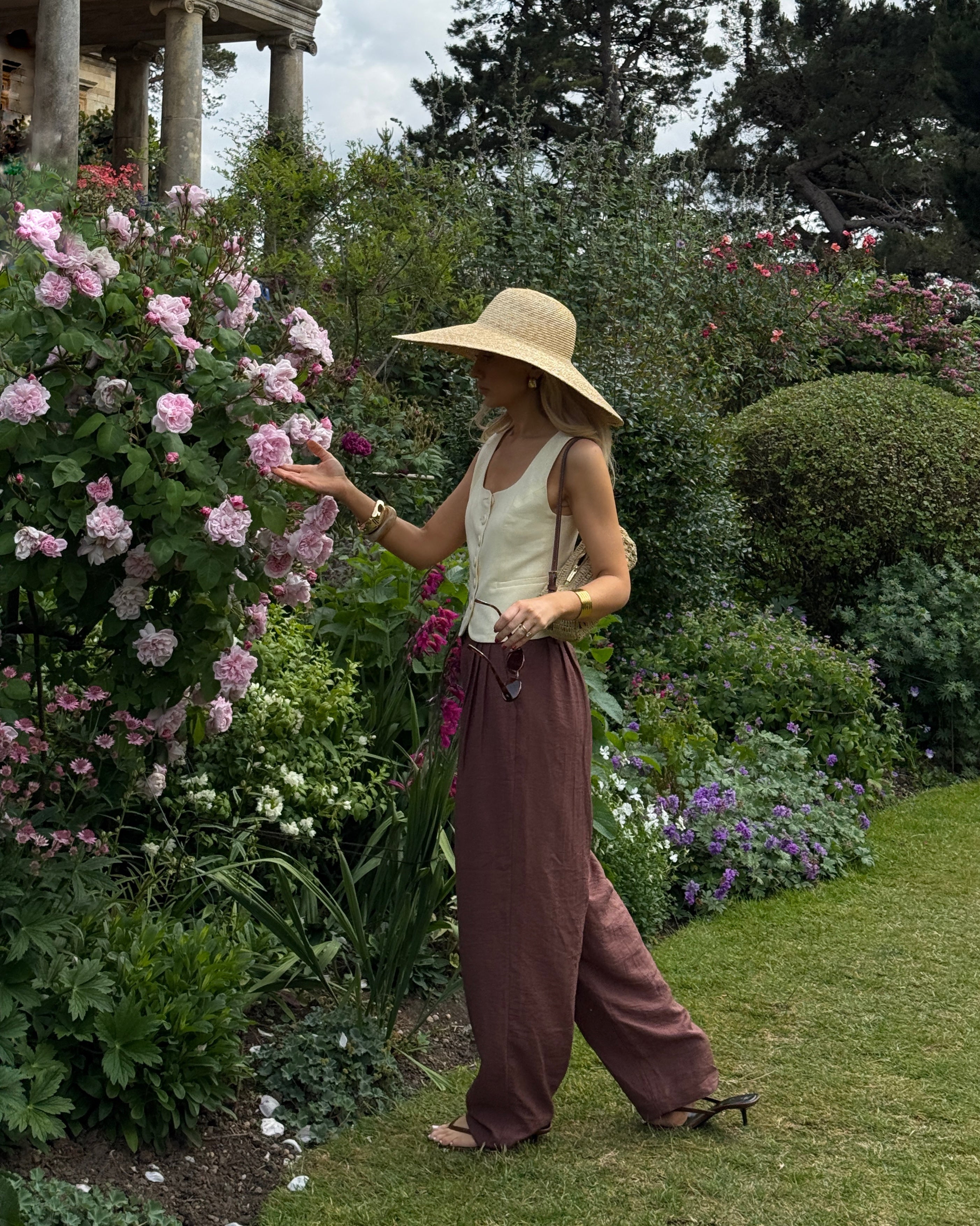 Woman in a vibrant flower garden wearing a wide-brimmed hat and flowing maroon trousers