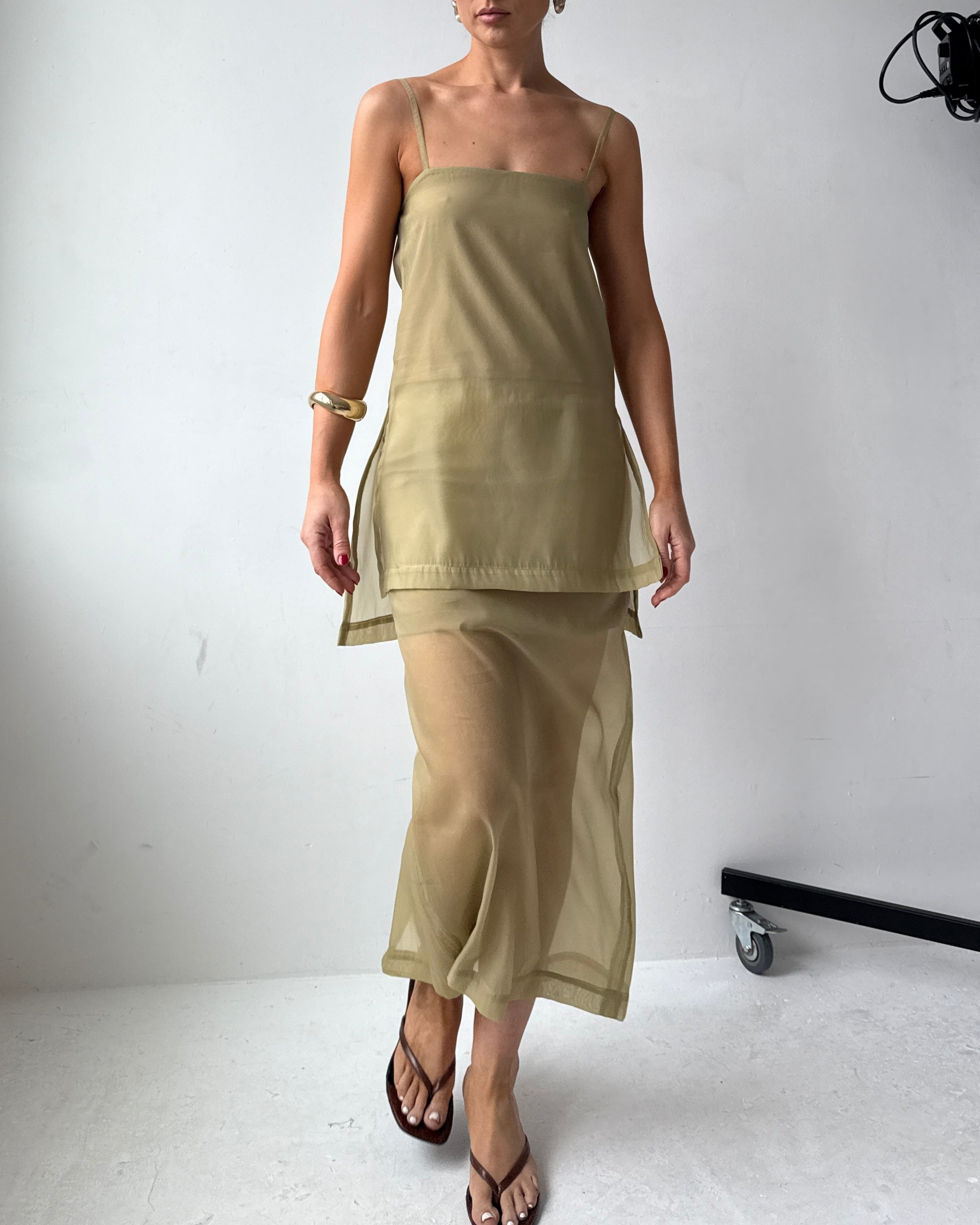 Model wearing a long light olive coloured semi-sheer top with thin straps and side slits up to the hips, paired with matching skirt and black sandals