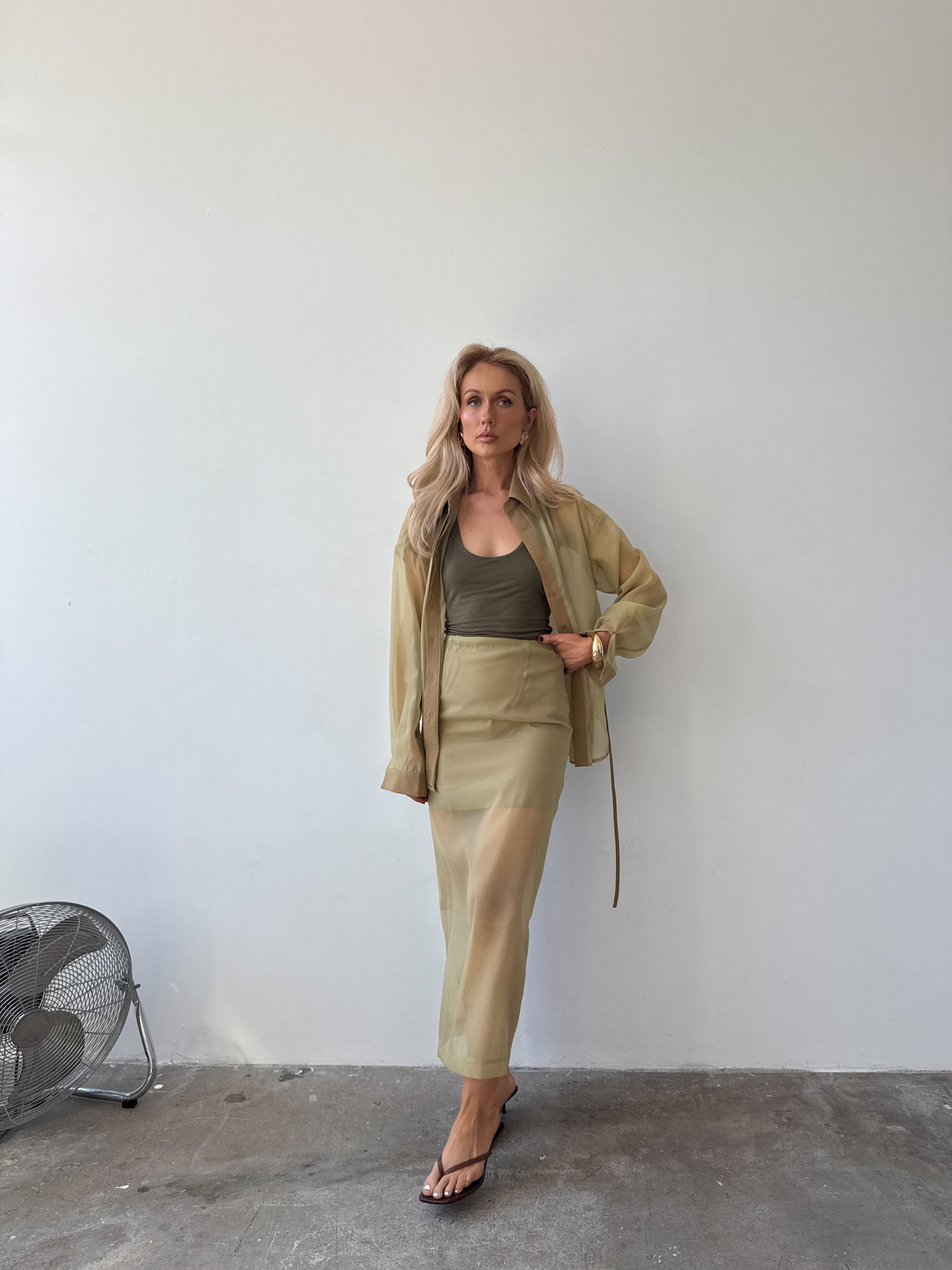 Model wearing a short, light olive opaque skirt layered under a longer sheer skirt, paired with a matching sheer shirt over an olive tank top, and sandals