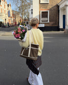 Woman crossing a street wearing a pale yellow top, holding flowers and carrying a brown and white bag