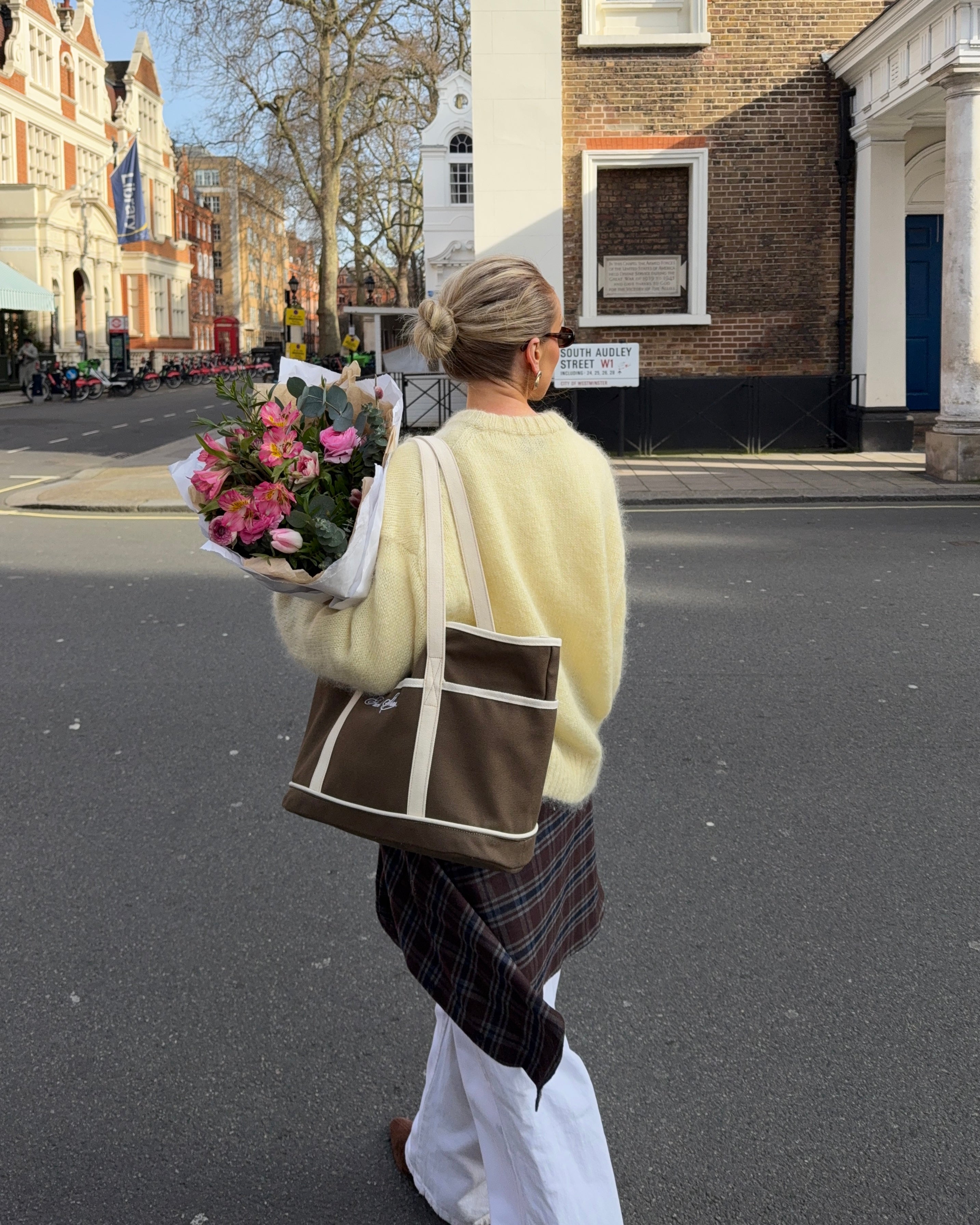 Woman crossing a street wearing a pale yellow top, holding flowers and carrying a brown and white bag