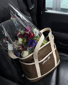 Brown and beige canvas tote bag with white text on the side, displayed in a car seat with a bouquet of flowers partially visible coming out of it.
