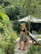 Model stands near a pool in a vibrant garden setting, wearing long checkered shorts, a black top, and a sun hat