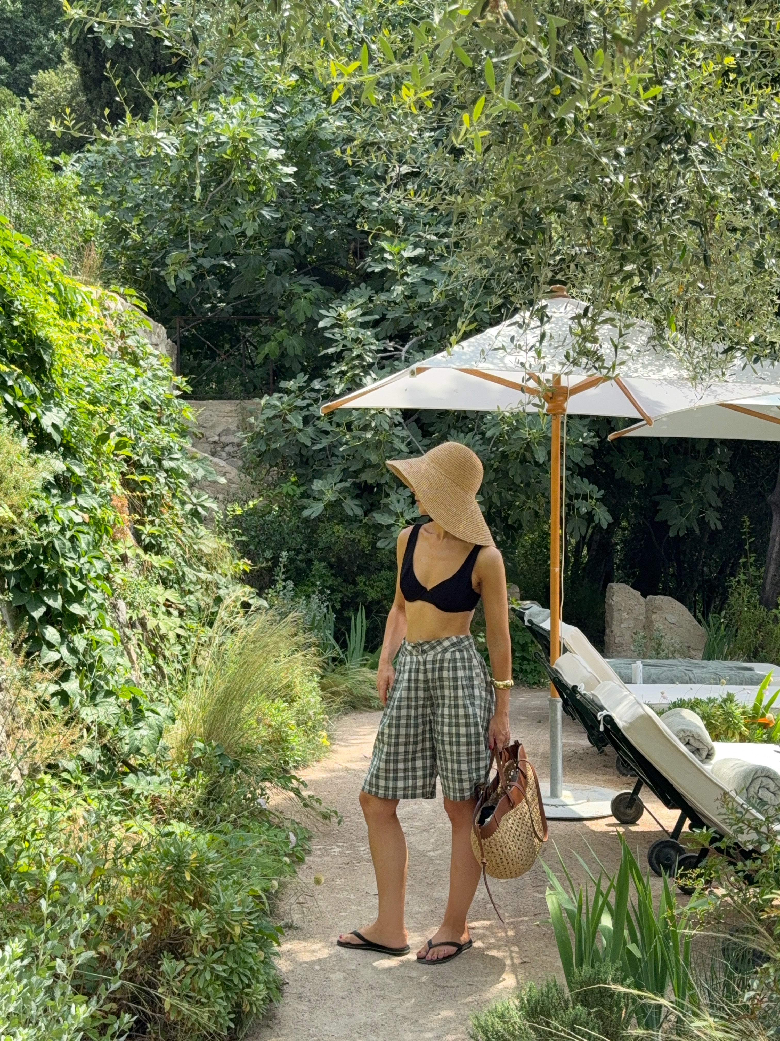 Model stands near a pool in a vibrant garden setting, wearing long checkered shorts, a black top, and a sun hat