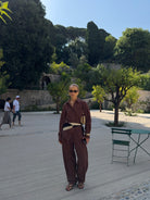 Woman in a brown outfit standing on a paved area with Mediterranean trees and walls in the background