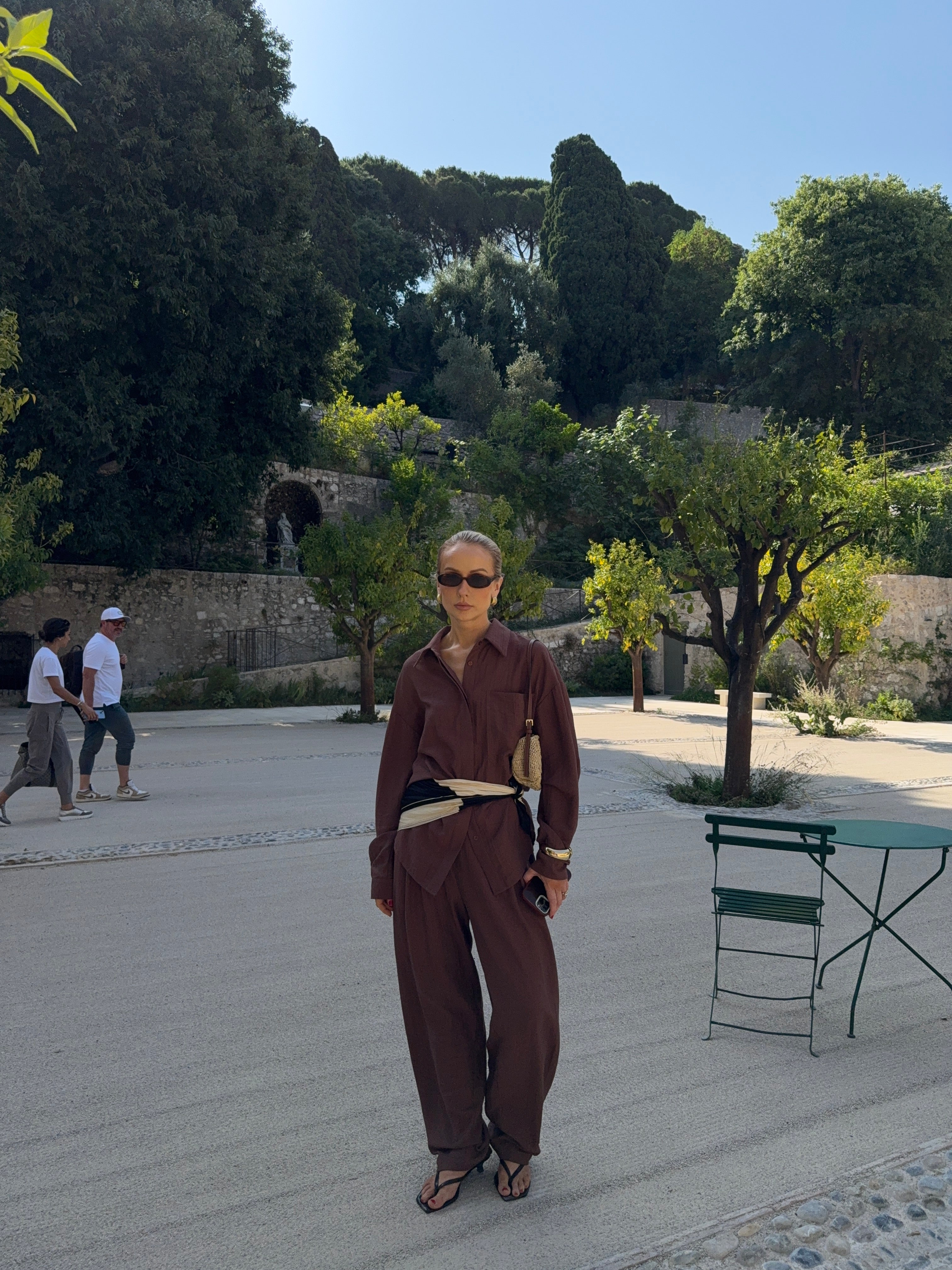 Woman in a brown outfit standing on a paved area with Mediterranean trees and walls in the background