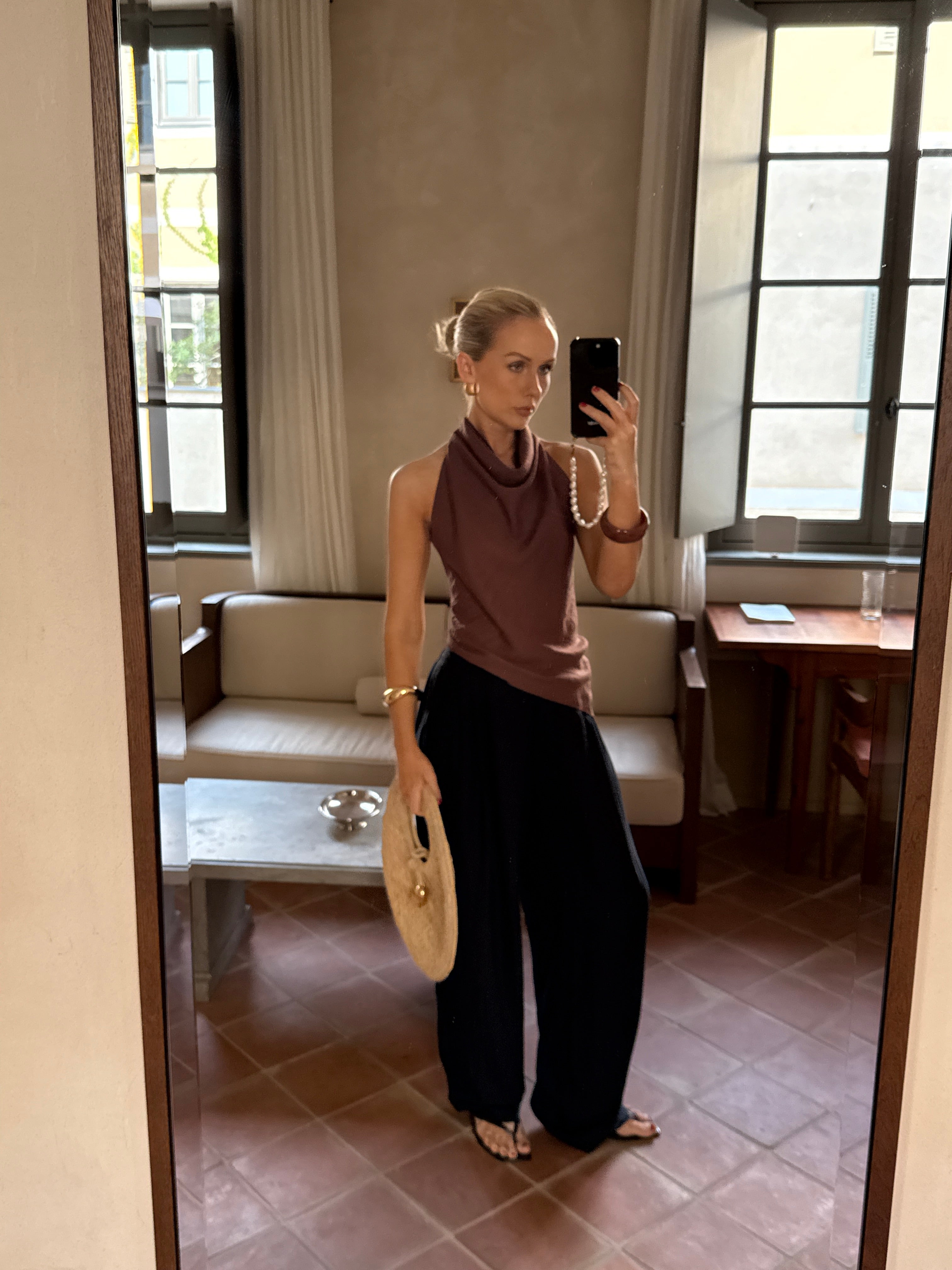 Model taking a mirror selfie wearing a brown top and black pants in a room with large windows
