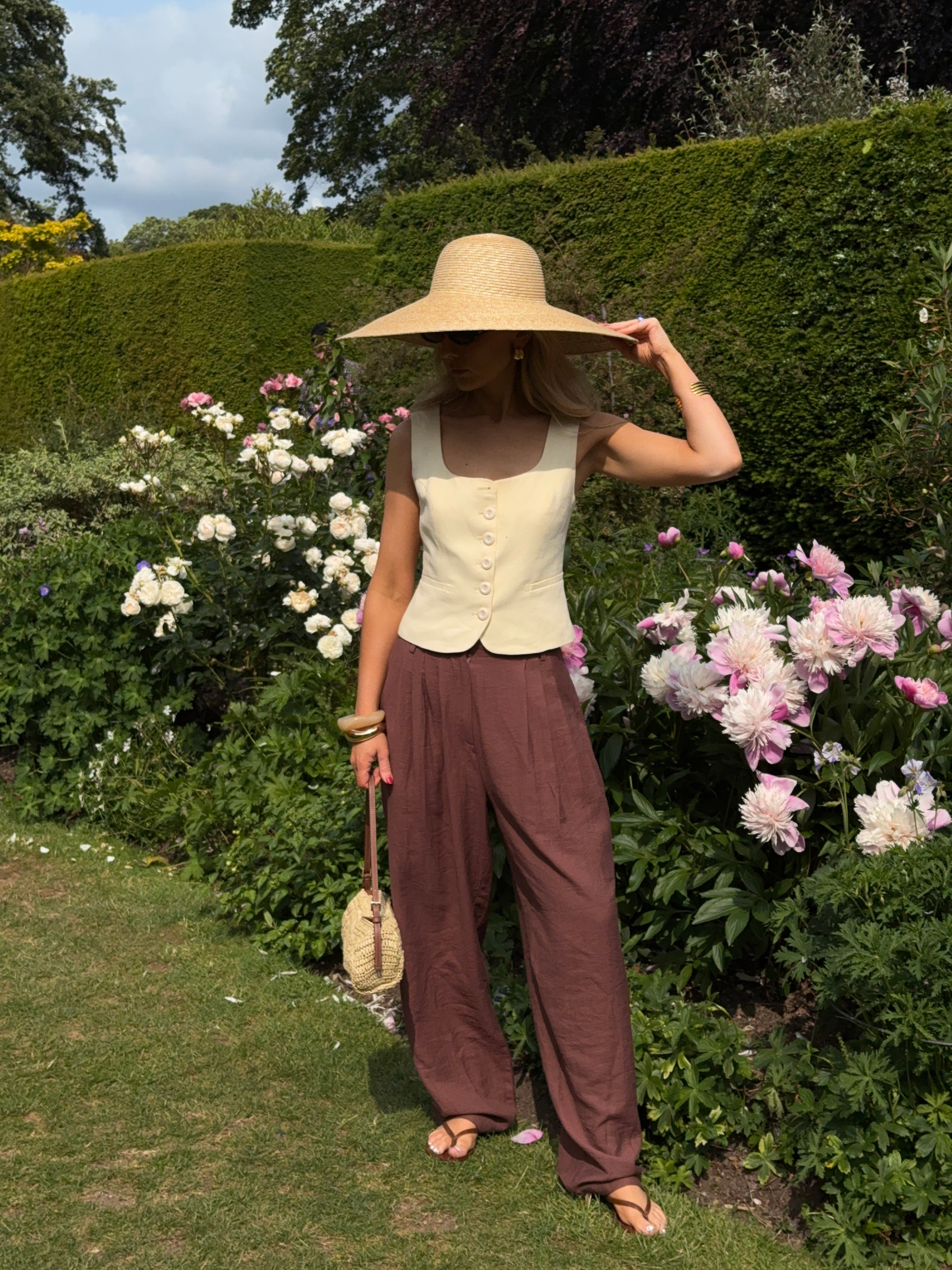 Woman in a sunny garden with box hedges and pale pink flowers flowers, wearing chocolate brown relaxed trousers, a pastel yellow waist coat, and a wide brimmed sunhat with matching handbag