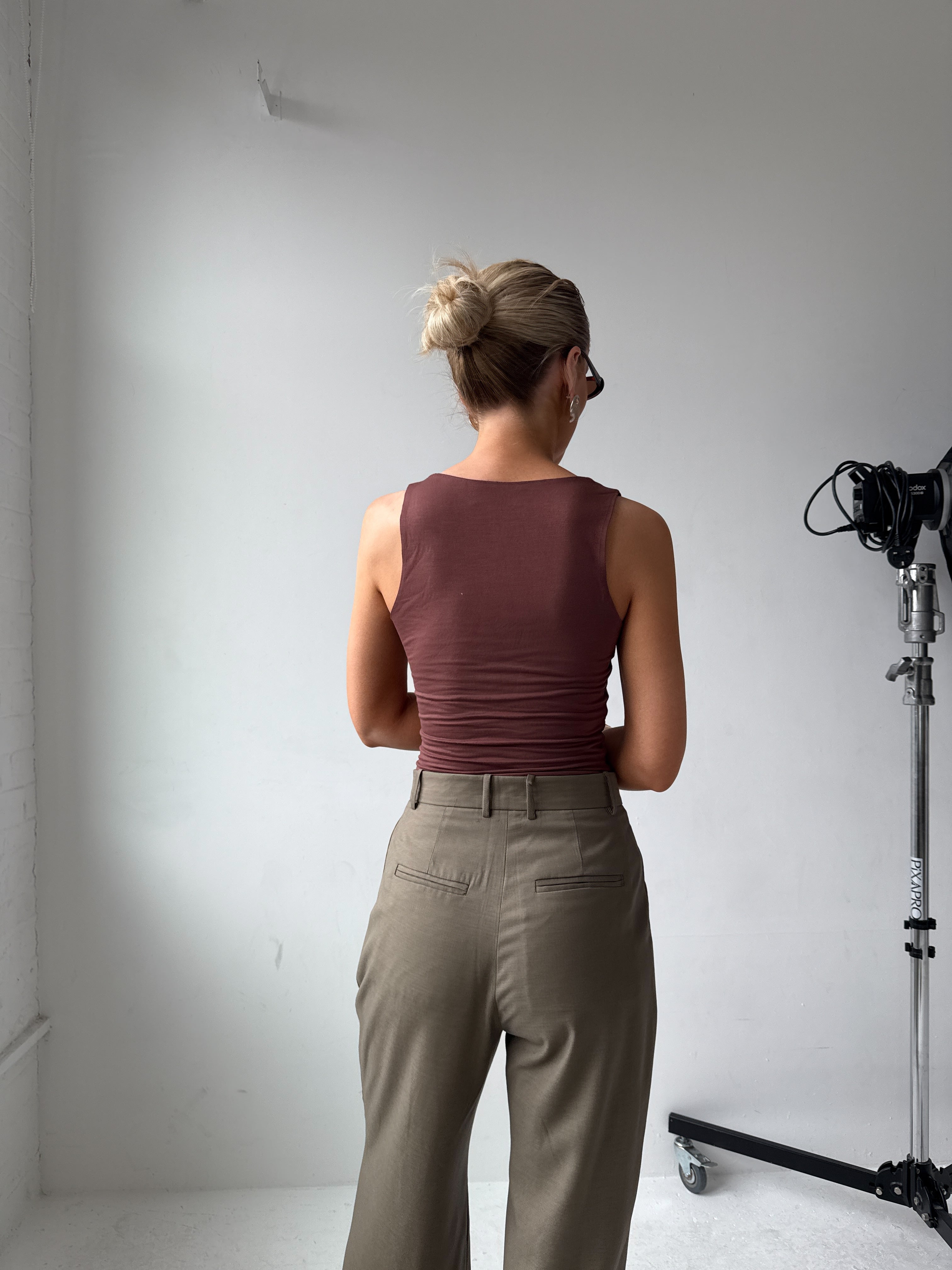 Model wearing a maroon top and beige-grey trousers standing in a studio setting with camera equipment