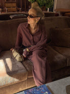 Model wearing a brown linen shirt with matching trousers on a brown sofa styled with sunglasses and a textured bag