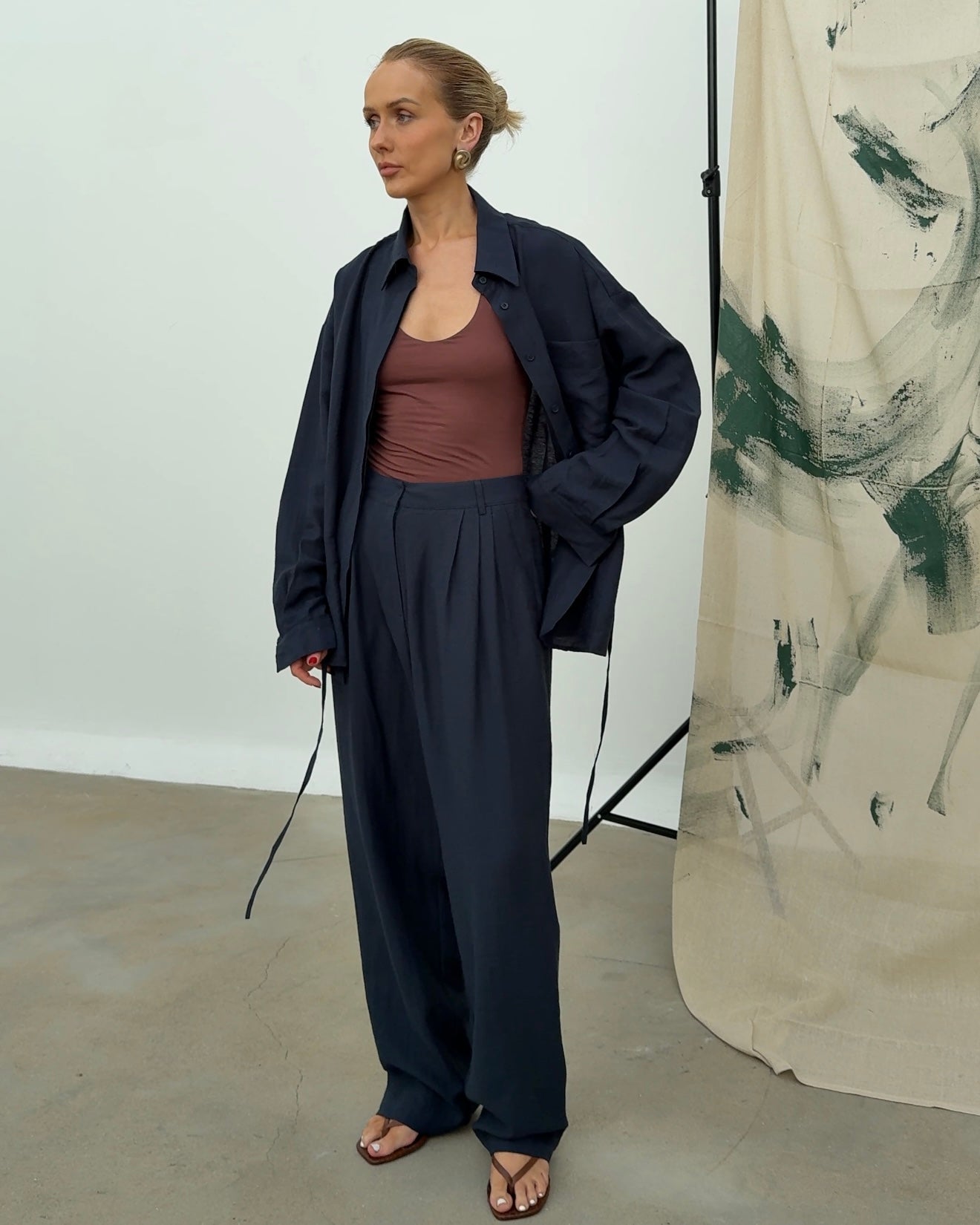 Model wears a navy linen shirt paired with matching trousers, standing in a minimalist room with an abstract painted background