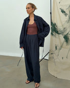 Model wears a navy linen shirt paired with matching trousers, standing in a minimalist room with an abstract painted background