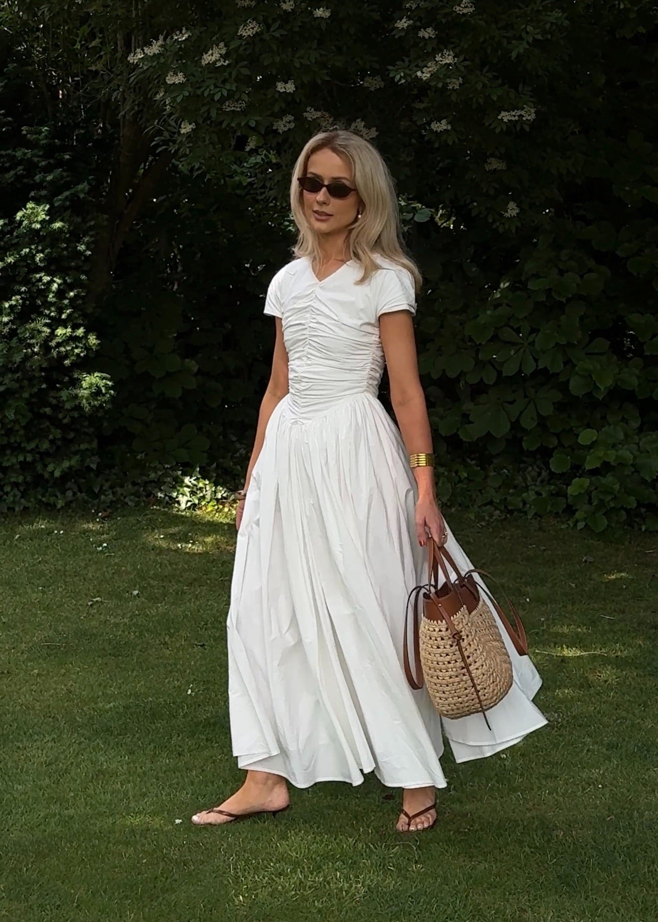 Model contrasting against an outdoor garden setting with a crisp white maxi dress featuring a v-neckline, short sleeves, full gathered fabric which is voluminous and flowing, styled with sunglasses, sandals and a straw tote